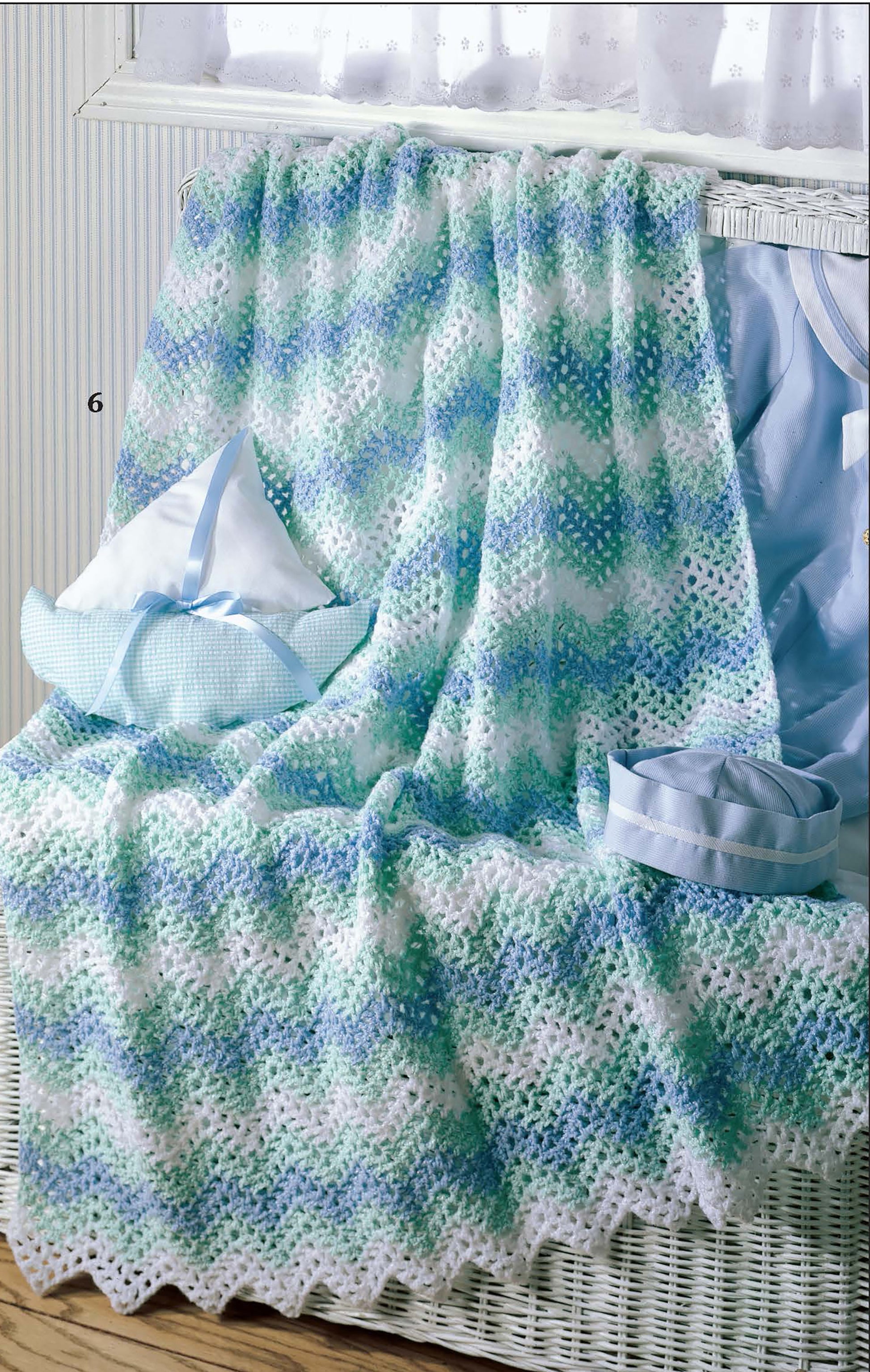 The "Ripples for Baby to Crochet" blanket by Leisure Arts features a soft, chevron pattern in white, blue, and green. It’s draped over a white wicker basket beside a light blue basket and a blue pillow.