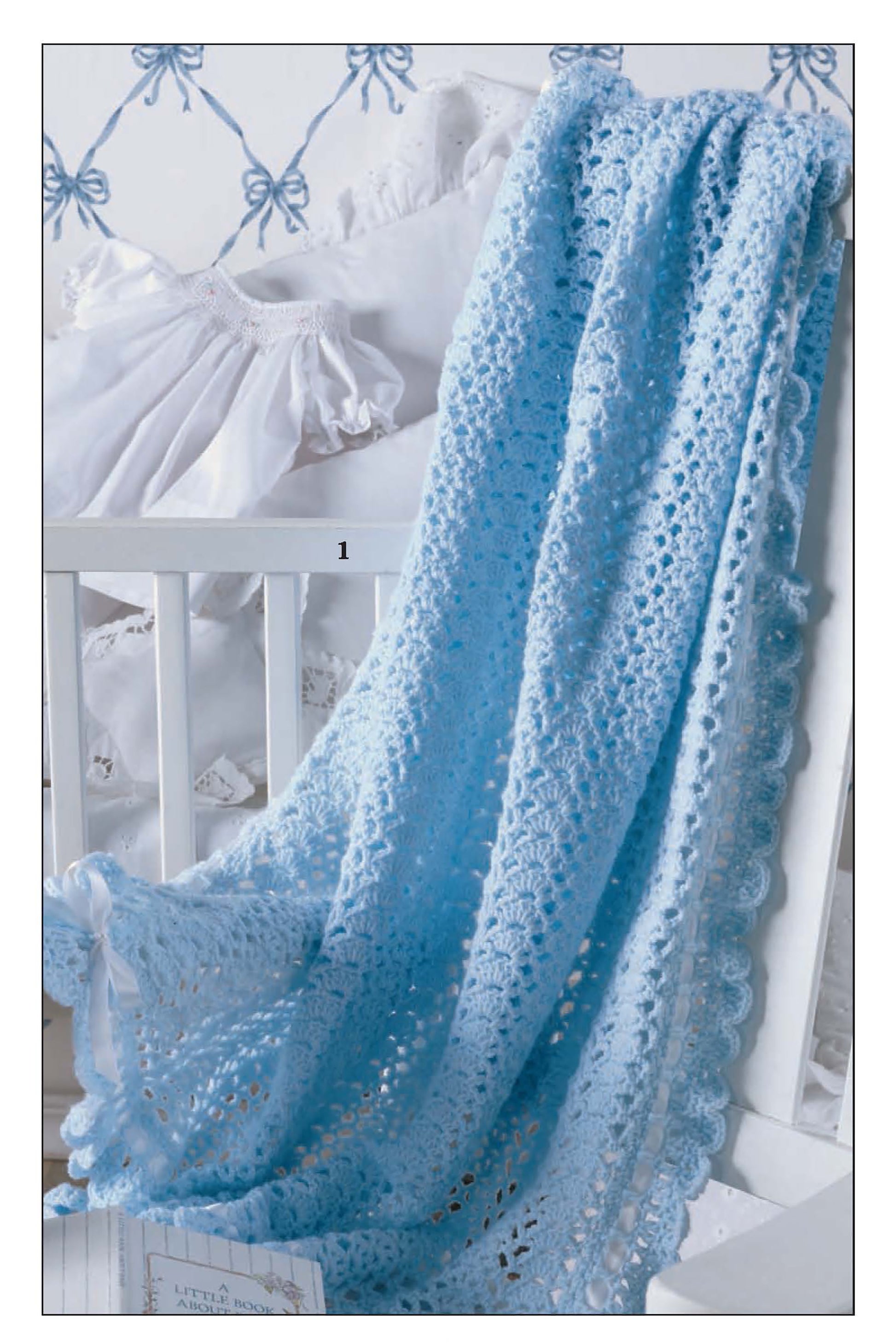 A light blue baby blanket from Leisure Arts' "So Sweet Baby Afghans" drapes over a white crib. White pillows and a ruffled baby dress sit behind it, with the wall displaying a blue bow pattern in the background.