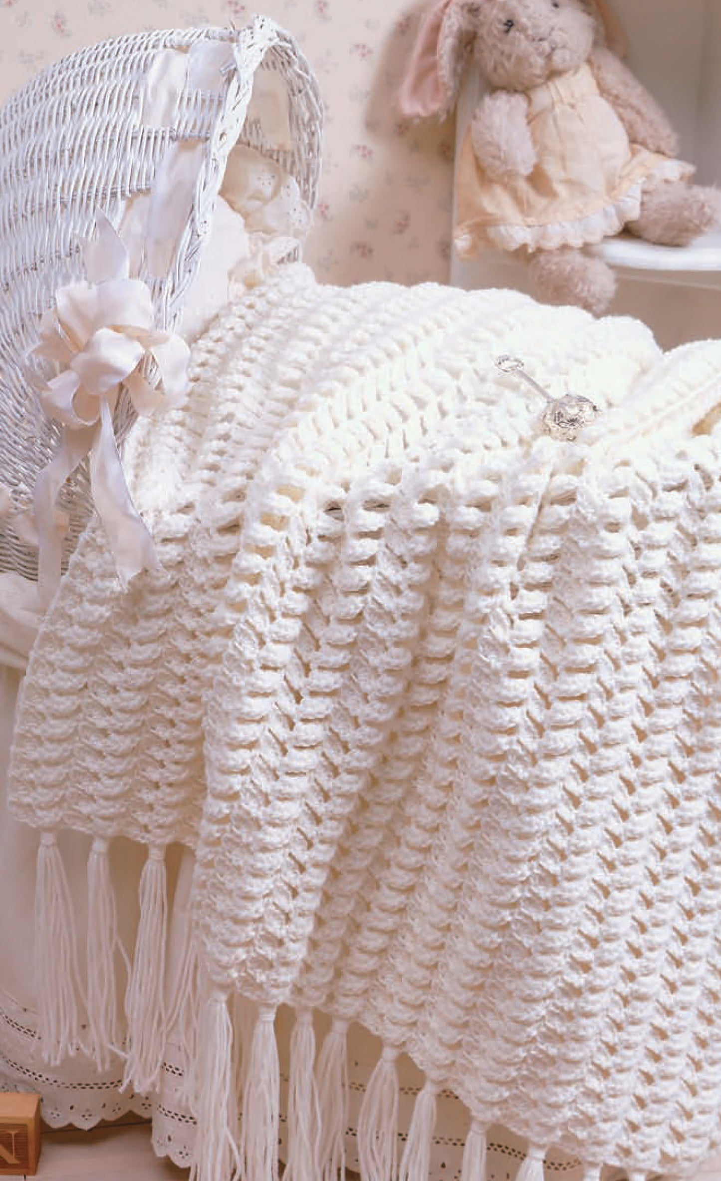 The “Nursery Favorites” by Leisure Arts features a white, crocheted worsted weight baby blanket with tassels draped over a wicker bassinet and a soft toy rabbit—perfect for thoughtful baby gifts and charming nursery décor.