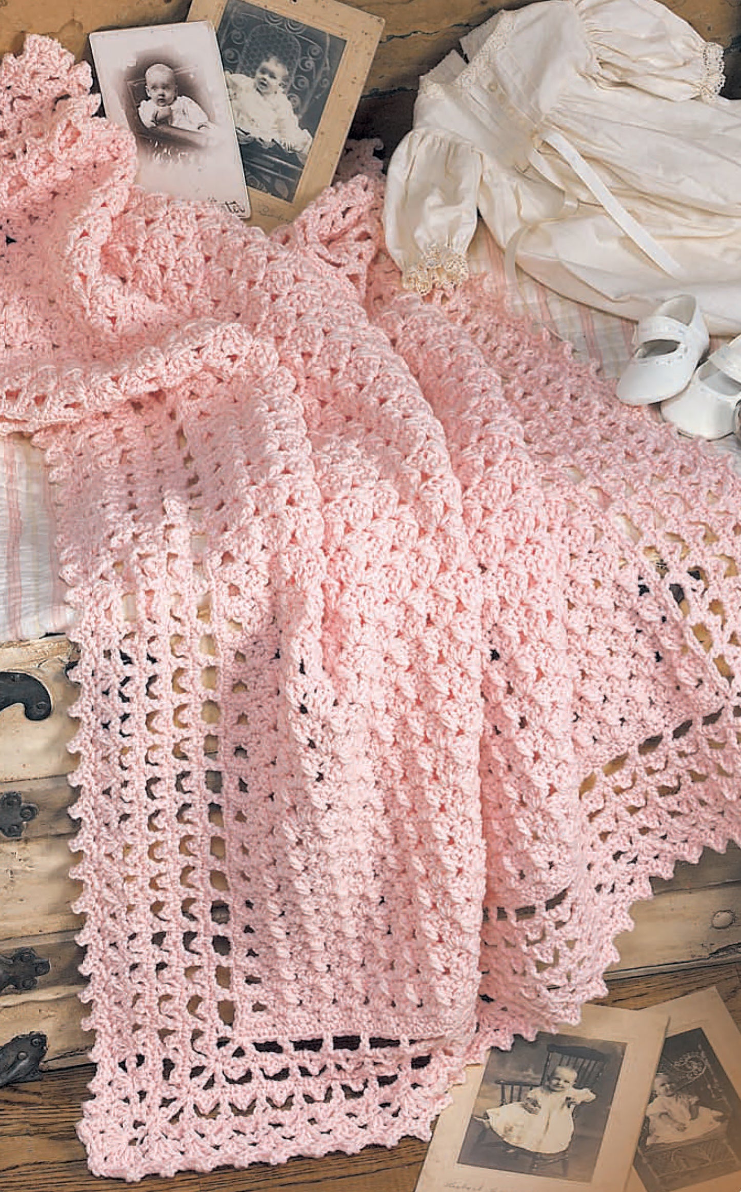 The Leisure Arts "Nursery Favorites" soft pink crocheted baby blanket is displayed with vintage baby photos, a white dress, and shoes, creating a nostalgic scene—a perfect treasured gift for any nursery.