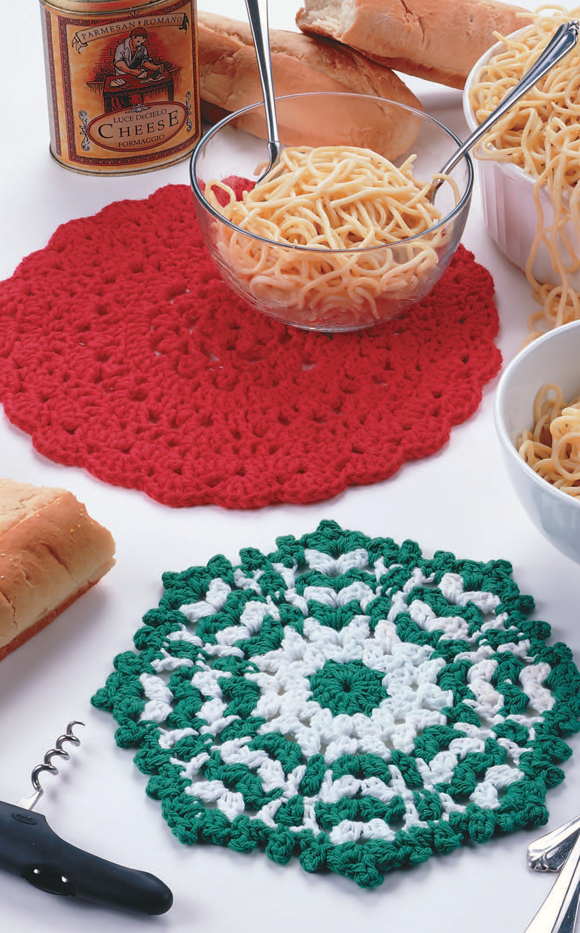 A glass bowl of spaghetti sits on a red placemat made with Little Kitchen Helpers by Leisure Arts, with another bowl nearby. A green and white placemat, baguette slices, a cheese tin, and corkscrew add to the cozy scene.