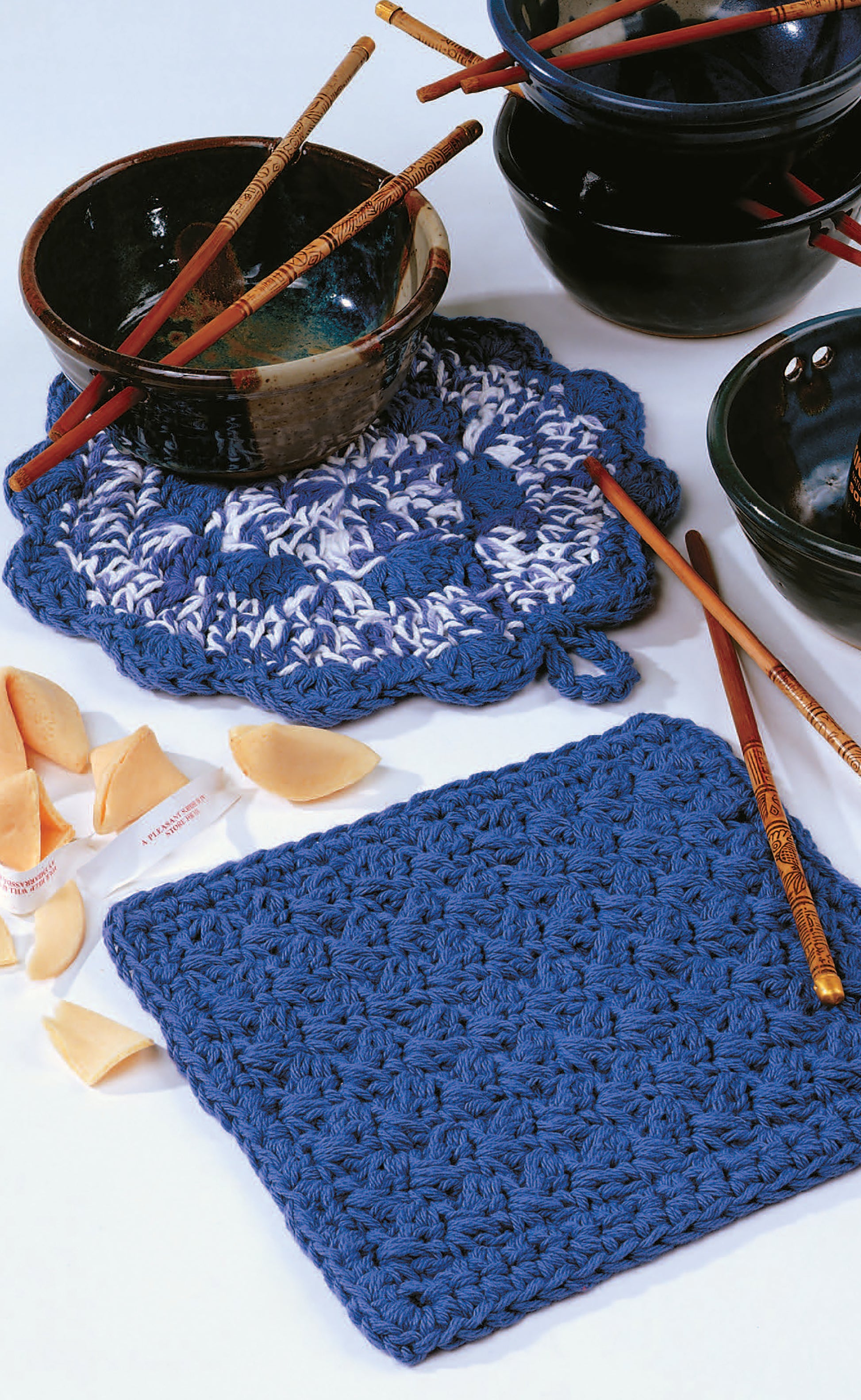 Add charm to your table with Little Kitchen Helpers by Leisure Arts: two blue crocheted pot holders—one solid, one scalloped—plus black bowls, chopsticks, and opened fortune cookies. Ideal as crochet gifts or for unique kitchen patterns.