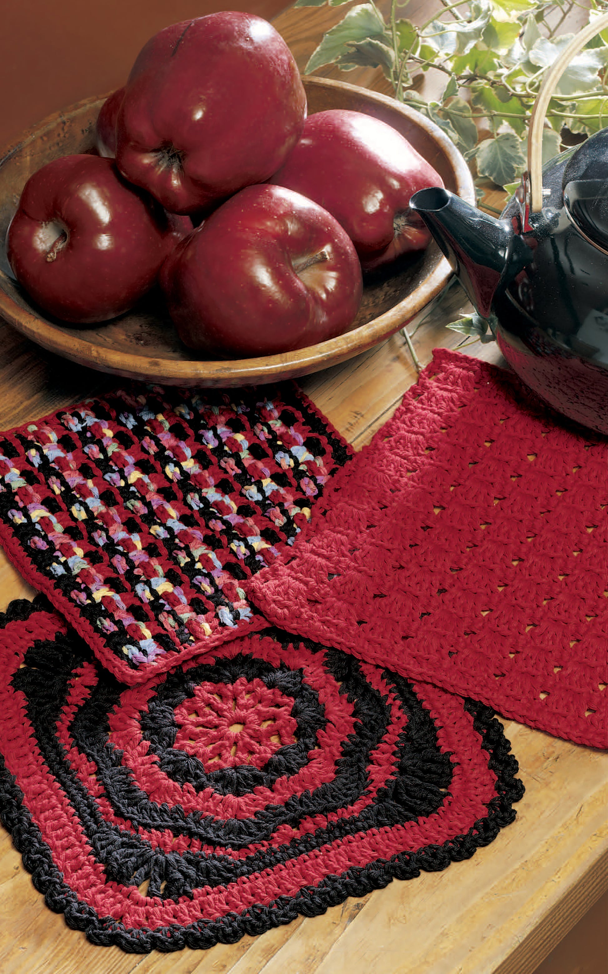 A wooden table displays Leisure Arts’ Little Kitchen Helpers—three crocheted pot holders in red, black, and multicolor—plus a bowl of red apples and a black teapot. Green ivy vines add to this perfect kitchen gift scene.