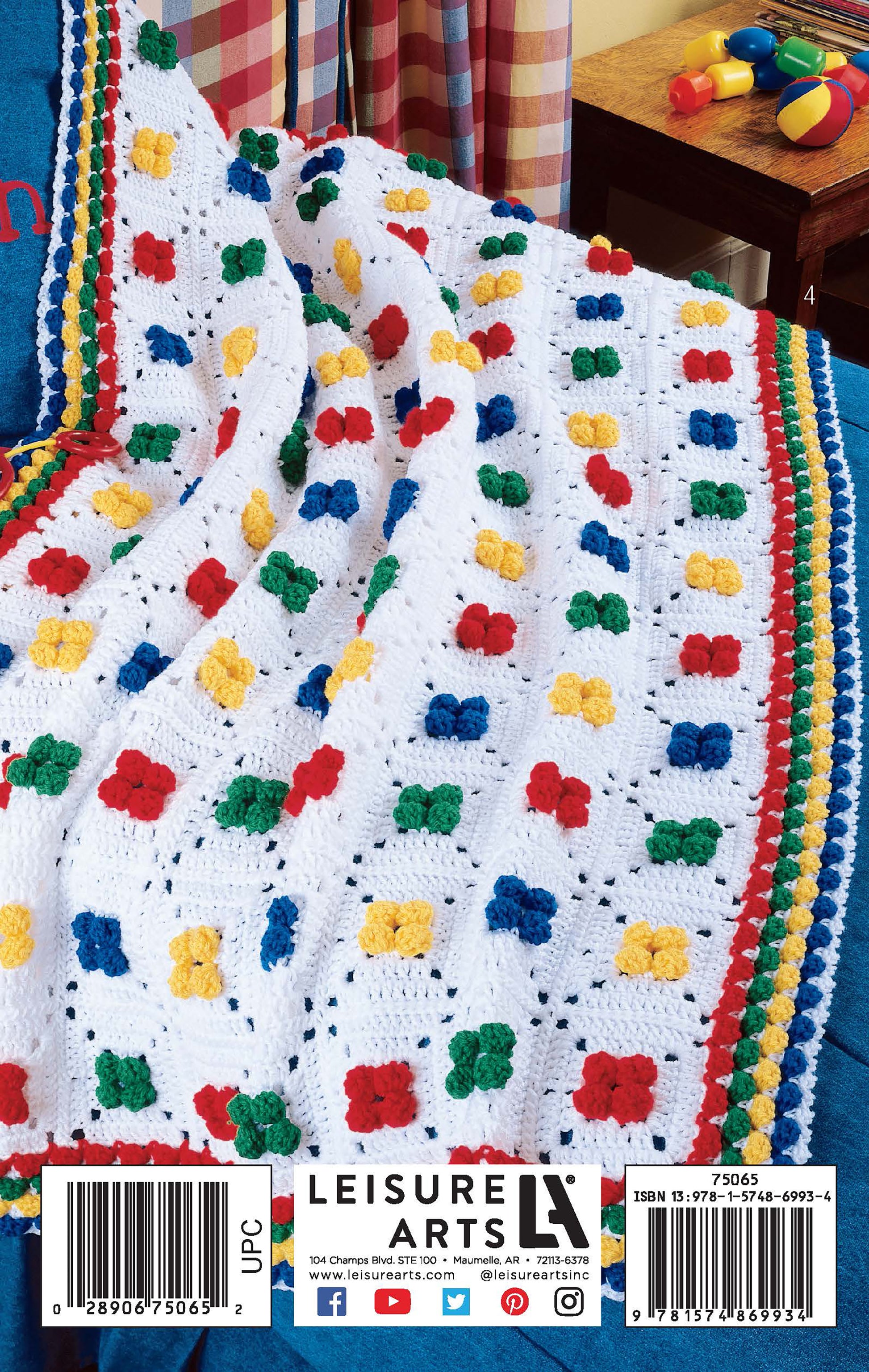 A cheerful crocheted blanket from the Kid-Pleasing Afghans collection by Leisure Arts, featuring colorful floral motifs, is displayed over a blue bedspread with toys and a plaid curtain in the background.