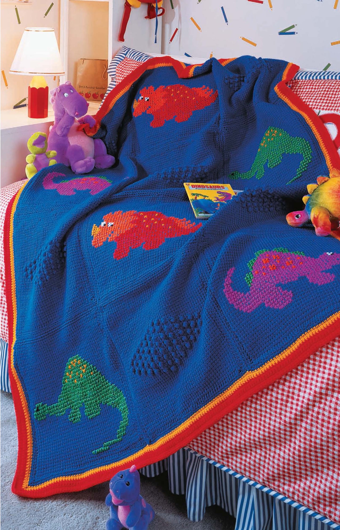 A cozy blue crocheted blanket from Leisure Arts' Kid-Pleasing Afghans, featuring vibrant dinosaur motifs, is draped over a bed with red gingham sheets. Plush dinosaur toys, a storybook, and a warmly glowing lamp complete the playful scene.