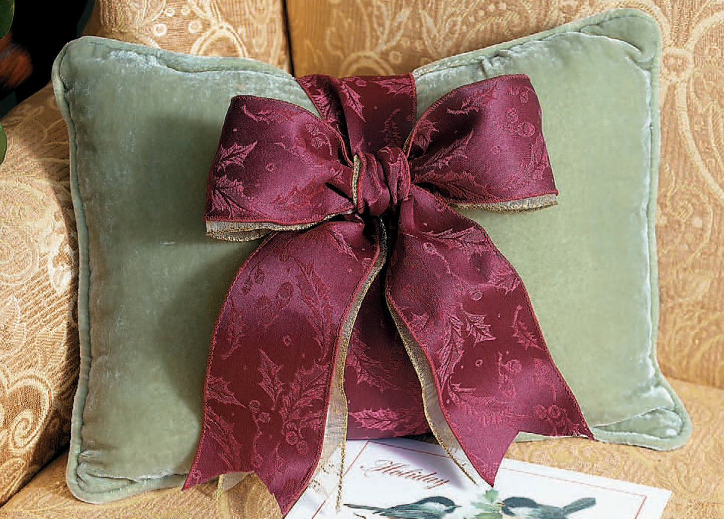 A patterned beige couch features The Little Book of Big Ribbon Ideas Digital Download from Leisure Arts, perfect for bow-making enthusiasts. A pale green velvet pillow with a large burgundy bow and a holiday card with a green tree are also visible.