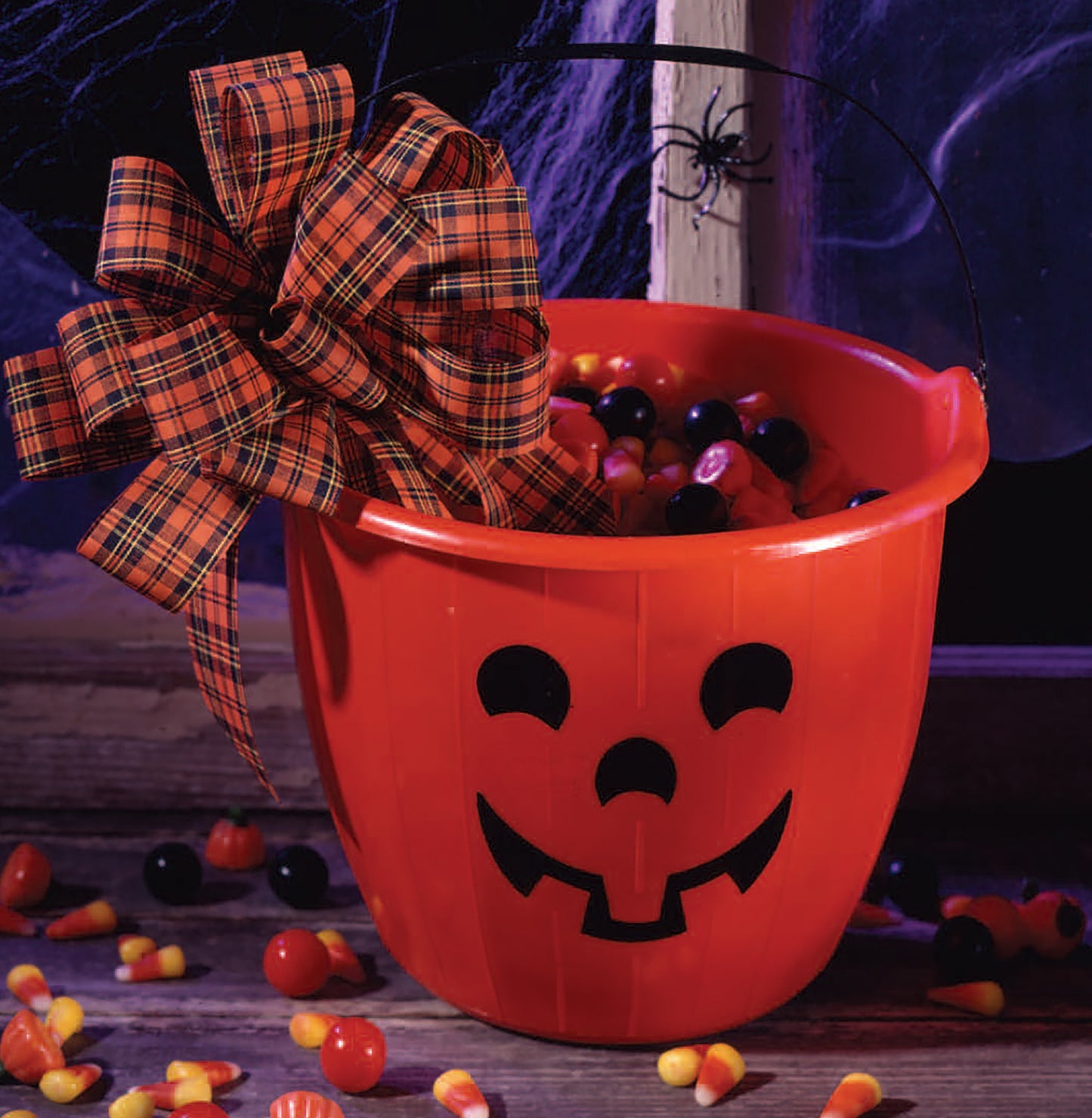 A wooden surface displays an orange Halloween bucket filled with candy corn and topped with a plaid bow, crafted using ideas from Leisure Arts’ The Little Book of Big Ribbon Ideas Digital Download. A spider decorates the handle, with scattered candies nearby.