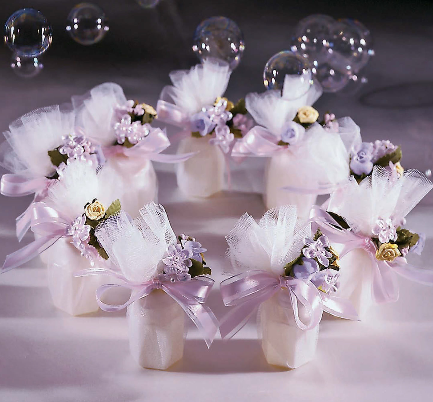 Six small white boxes decorated with pastel flowers, tulle, and pale pink ribbons from The Little Book of Big Ribbon Ideas by Leisure Arts are arranged in a circle on purple, with soap bubbles floating above as charming gift toppers.