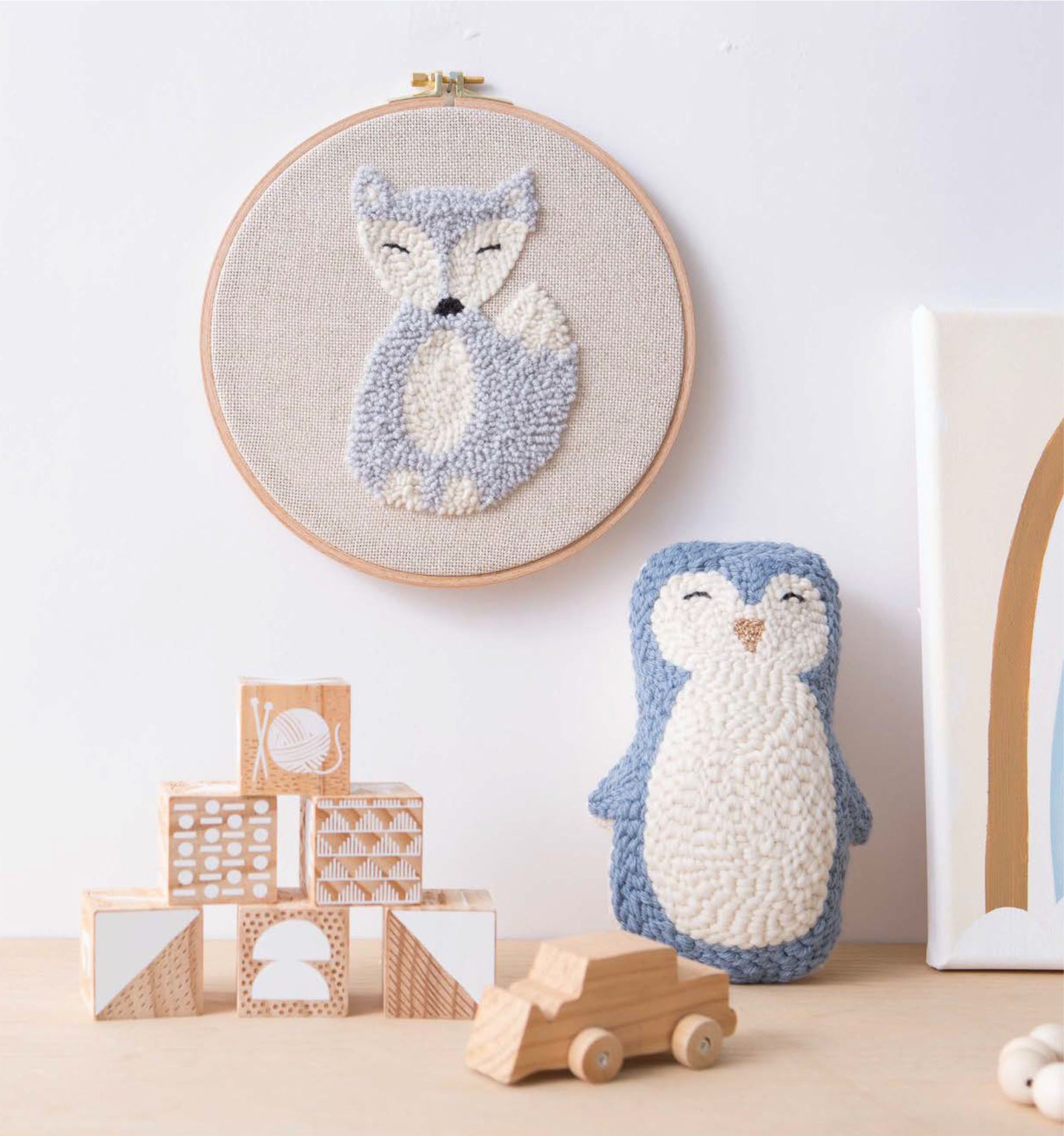 A display features the Leisure Arts Modern Punch Needle fox embroidery hoop art, a handmade plush penguin, assorted wooden blocks, and a small wooden toy car on a light wooden surface.