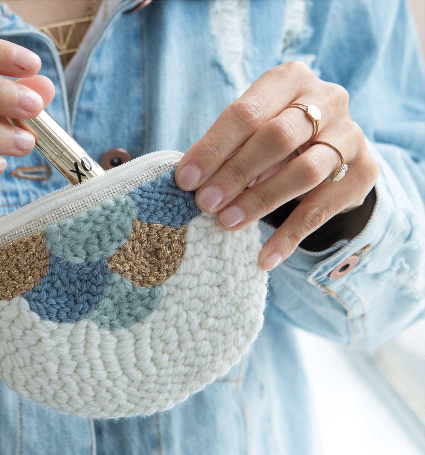 Wearing a denim jacket, a person holds the Leisure Arts Modern Punch Needle pouch with pastel crochet patterns—ideal for lipstick or storing small punch needle projects. Gold rings and manicured nails complete this stylish, crafty scene.