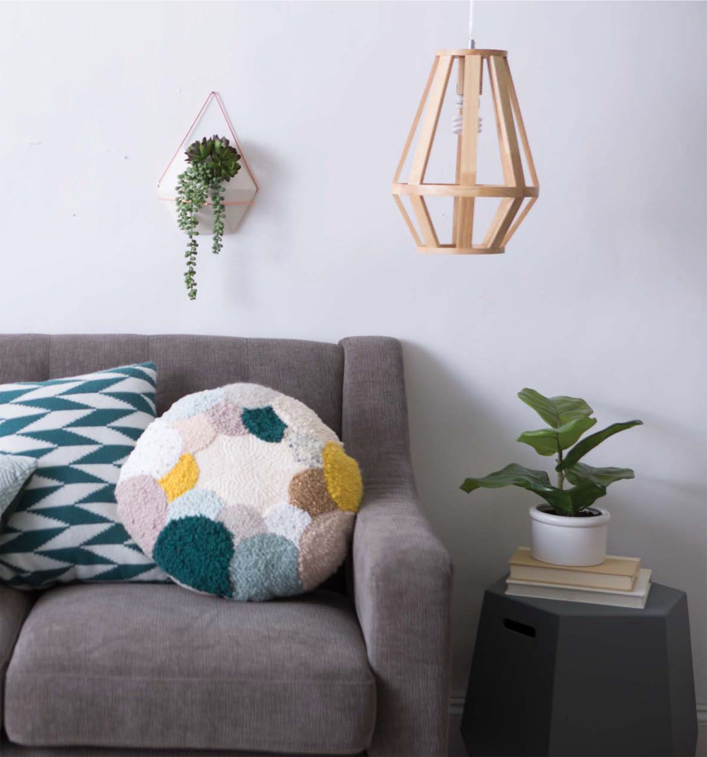 A modern living room with a gray sofa, textured cushions inspired by Leisure Arts’ Modern Punch Needle, a hanging wooden light fixture, a potted plant on a gray side table, and a wall planter with trailing greenery.