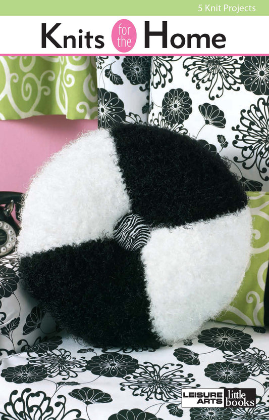 The book "Knits for the Home" by Leisure Arts is shown on a bed with black and white floral bedding, featuring a round, fuzzy black and white pillow with a patterned button center as stylish knitted home decor.