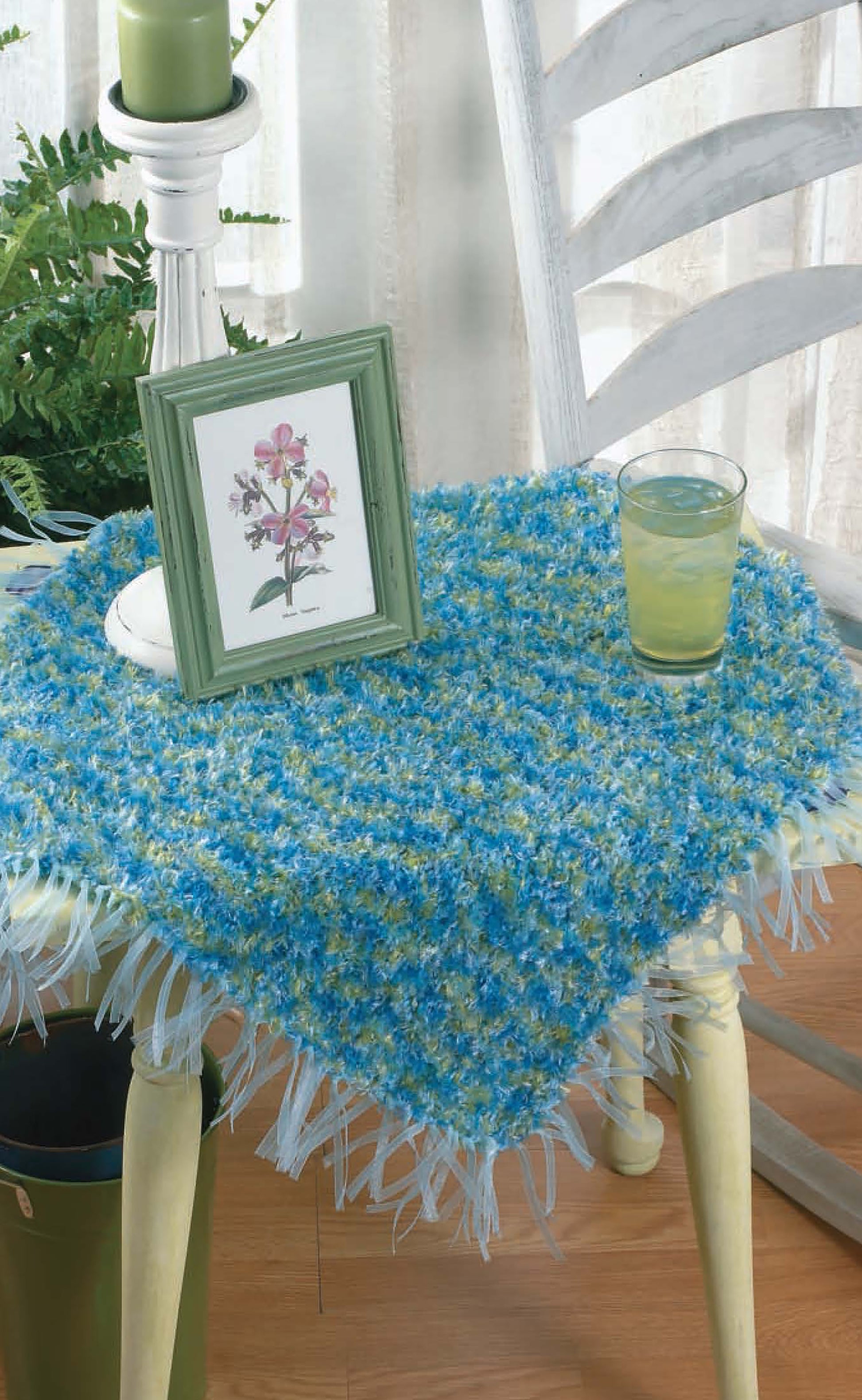 A small table with a blue and green textured, fringed cloth—ideal inspiration for Knits for the Home by Leisure Arts—displays a green candle, framed botanical art, and lemonade. Nearby sit a white chair and a houseplant.
