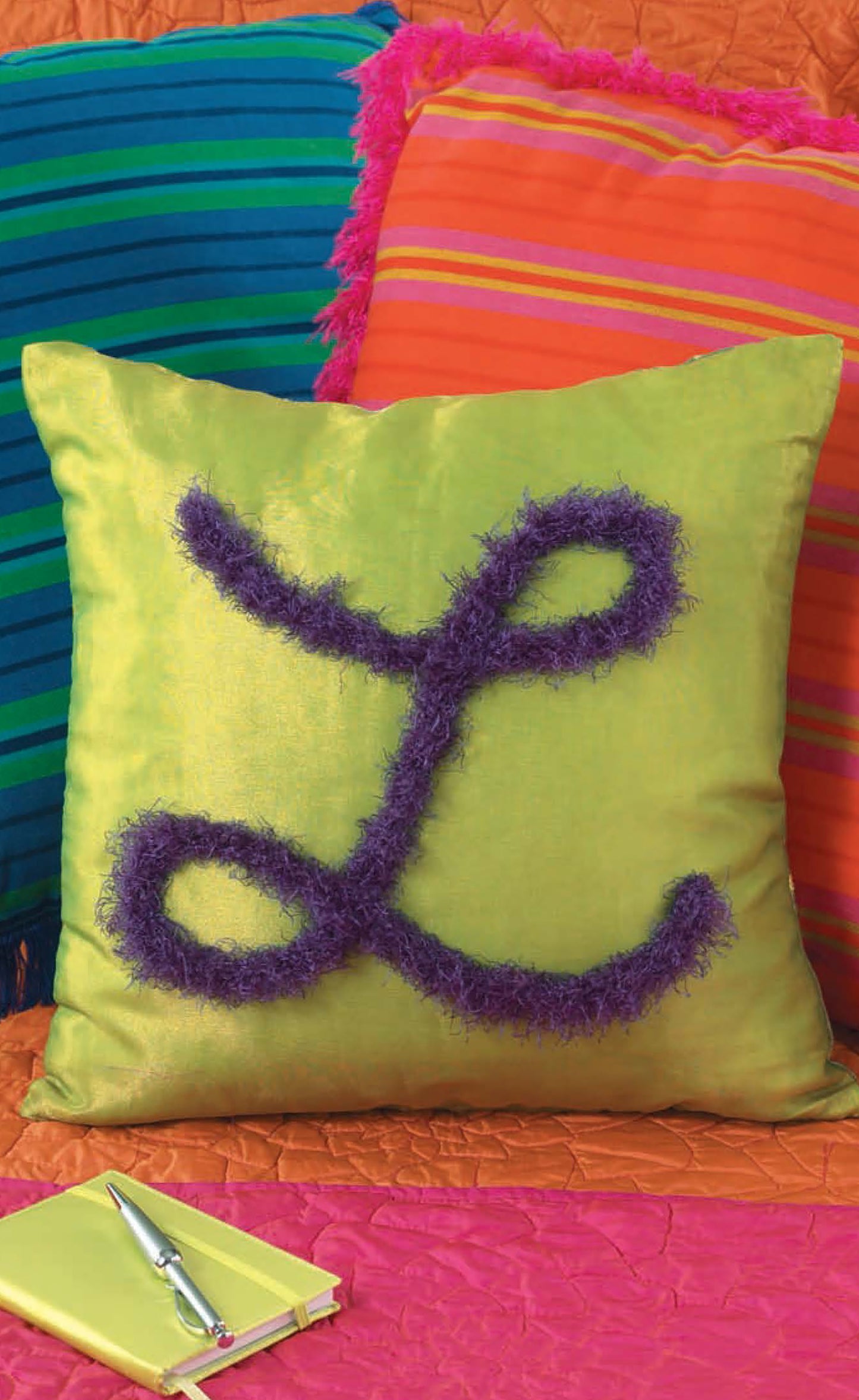 A green pillow with a large purple fuzzy "L," inspired by beginner projects in Knits for the Home by Leisure Arts, sits before striped and orange pillows. A yellow notebook and silver pen rest on a pink surface nearby.