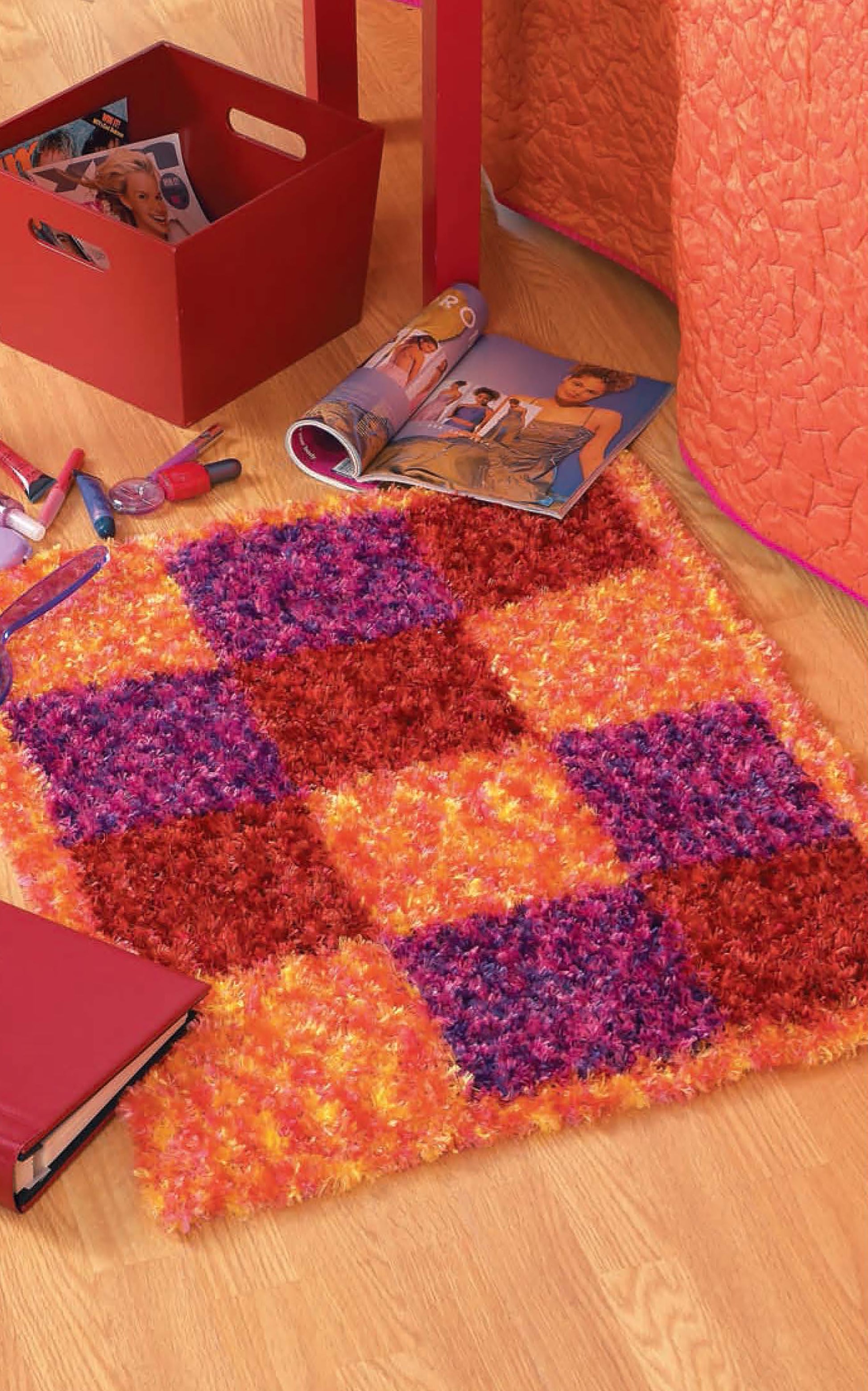 The Leisure Arts Modern Punch Needle shaggy rug showcases red, orange, and purple checkerboard squares beside an orange bed, open magazines, toys, and a red storage box on a wooden floor.