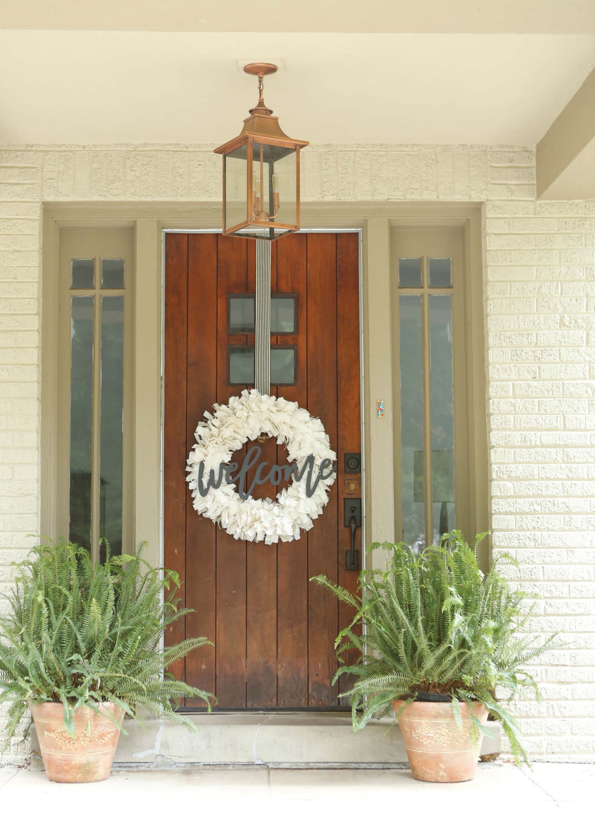 The wooden door displays a white “Welcome” wreath, offering inspiration for beginner DIYers using the Quick Wreath Designs Digital Download by Leisure Arts. Two potted ferns and a lantern-style fixture complete the entryway.