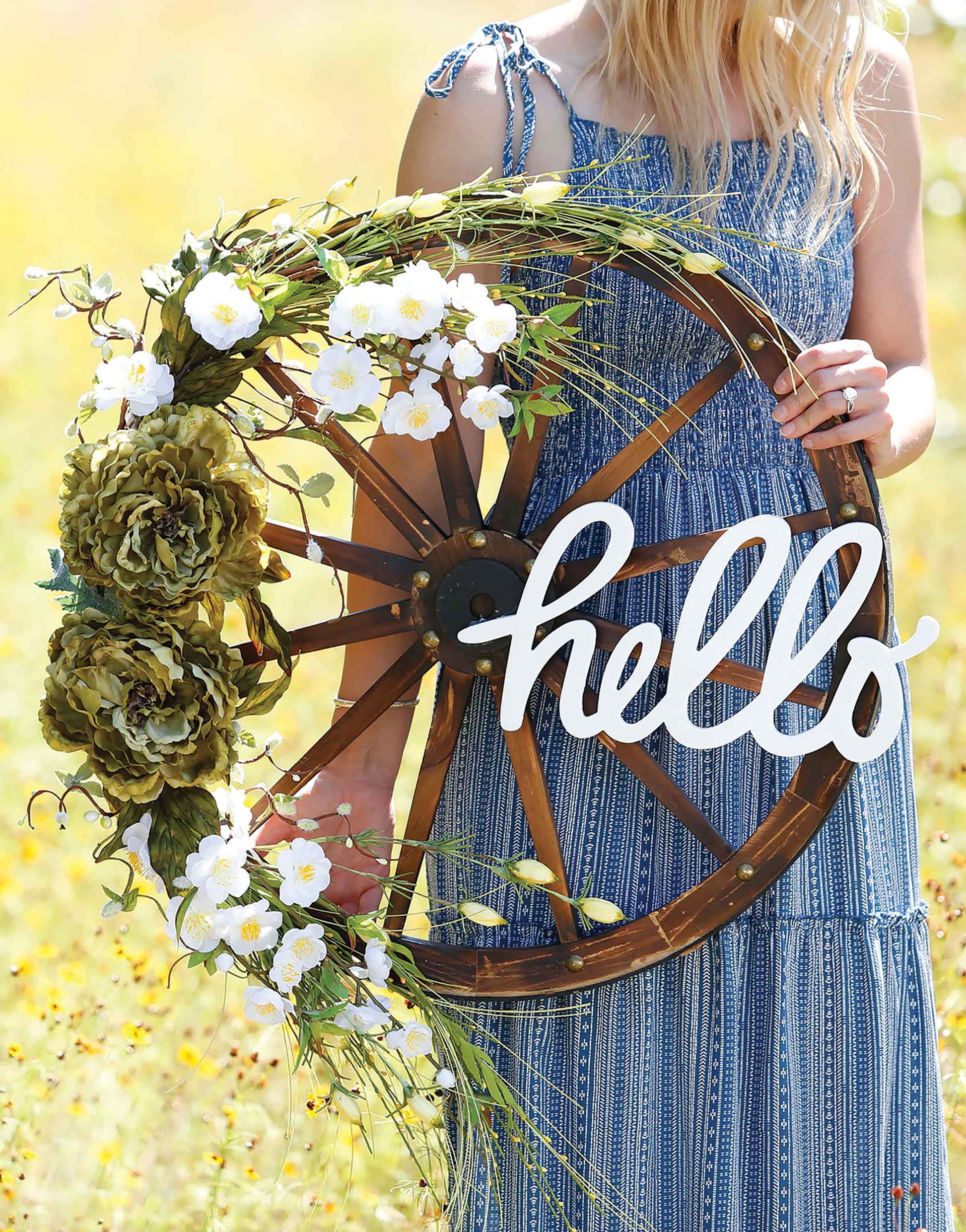 A woman in a blue patterned dress showcases the Quick Wreath Designs Digital Download by Leisure Arts—holding a rustic wagon wheel adorned with green and white flowers plus a “hello” sign—perfect inspiration for beginner DIY wreath projects.