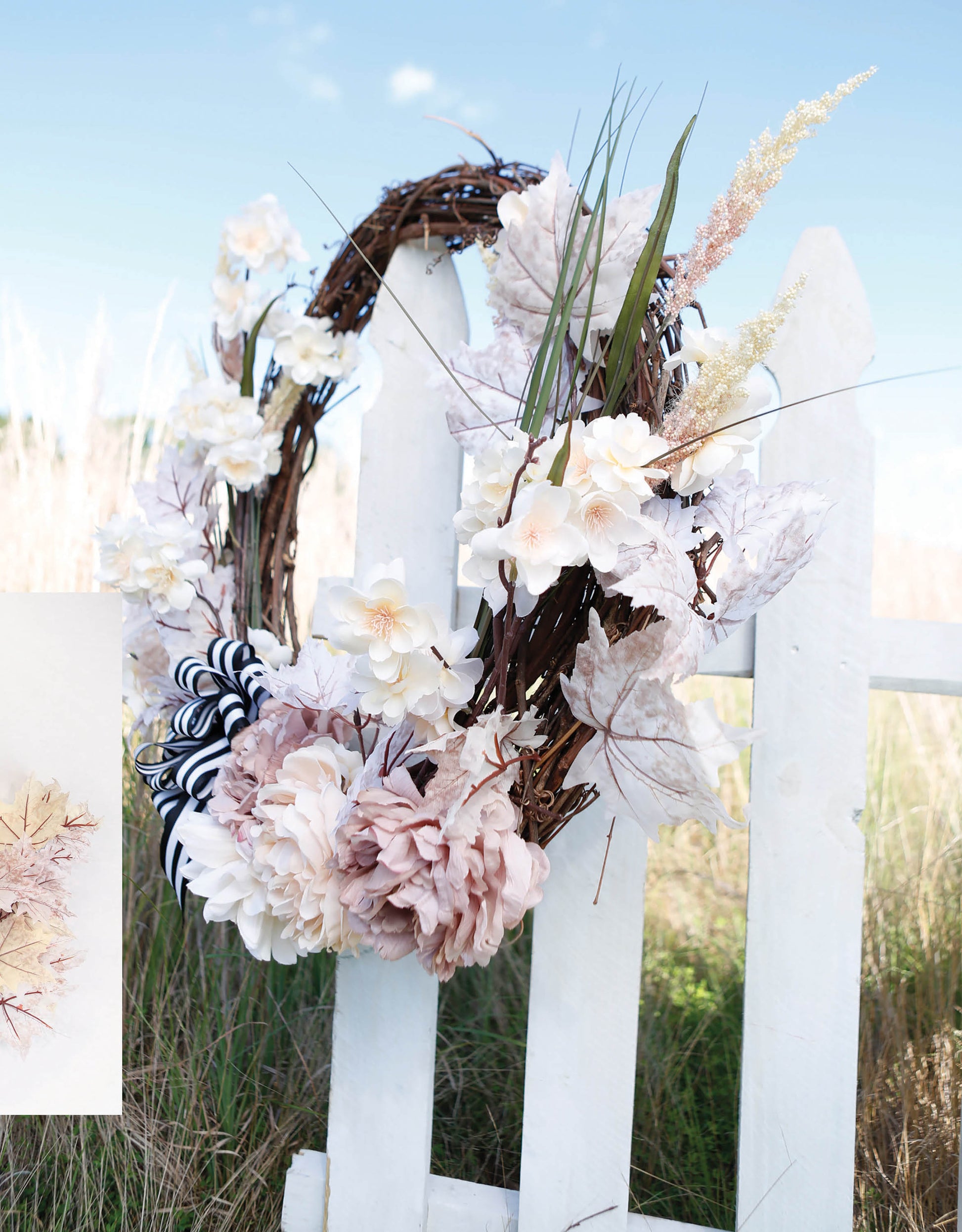The Quick Wreath Designs Digital Download by Leisure Arts features a white and pale pink floral wreath hanging on a white wooden fence, perfect for home decor craft inspiration with tall grass and blue sky in the background.