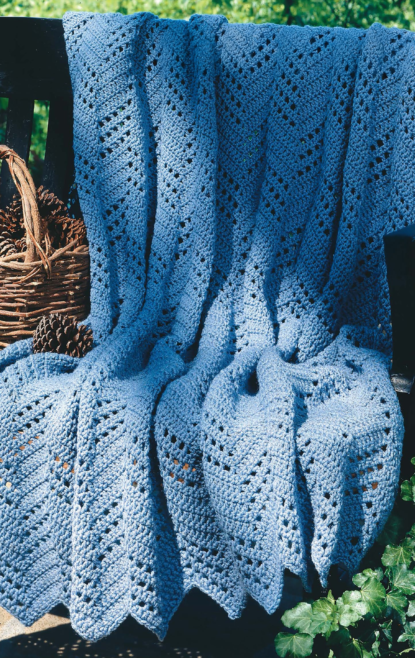 A digital guide from Leisure Arts, "Beginner's Guide Ripple Afghans," features chevron lace knit patterns like the light blue blanket draped outdoors on a wooden bench, beside a wicker basket of pinecones and lush green foliage.