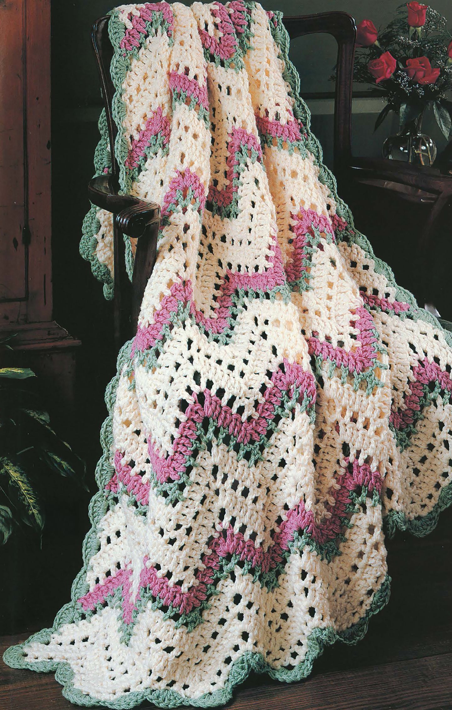 The Beginner's Guide Ripple Afghans Digital Download by Leisure Arts features cream, pink, and green ripple crochet blanket patterns, as shown draped over a dark wooden chair with a vase of red roses in the background.