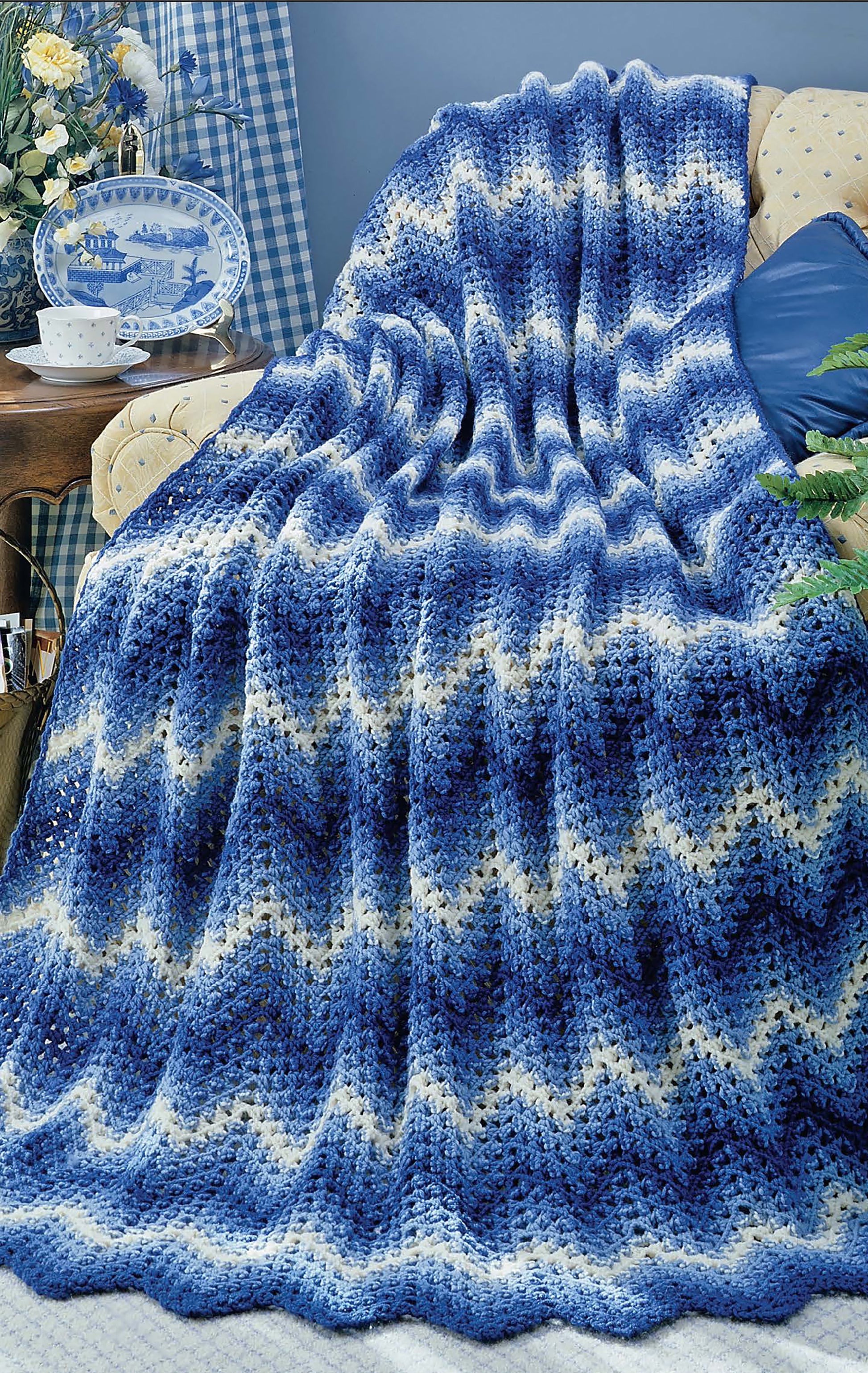The Leisure Arts Beginner's Guide Ripple Afghans Digital Download features a blue, white, and navy chevron crochet blanket draped over a tan armchair in a cozy room with checkered fabric, a blue-and-white plate, and plants.