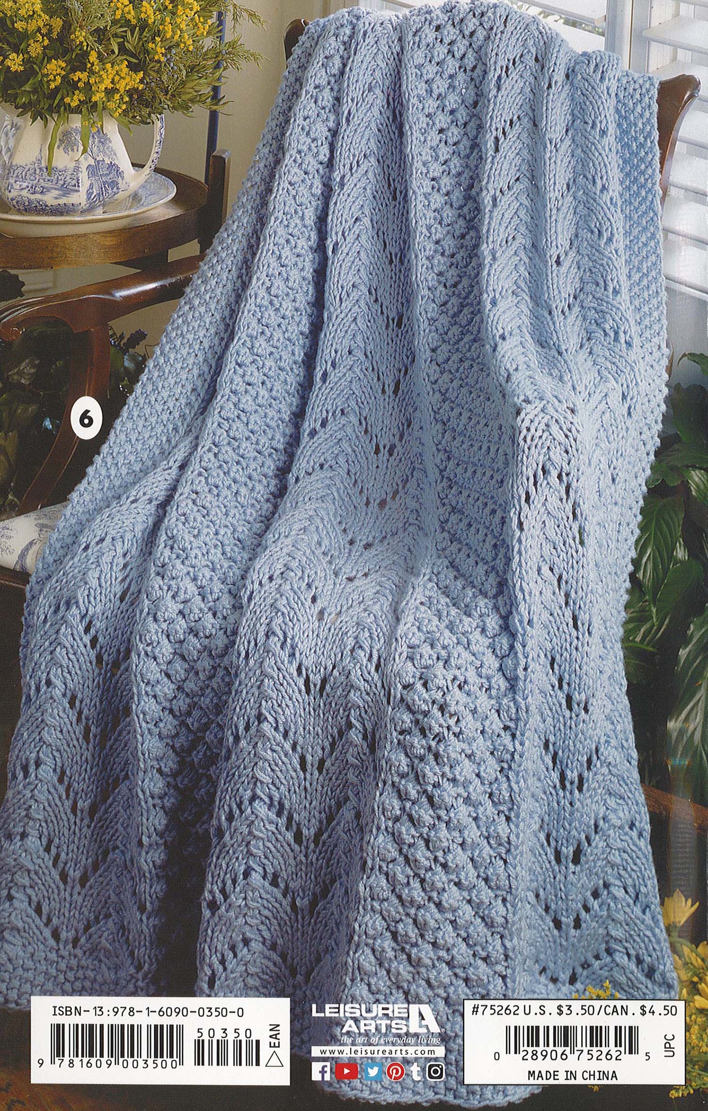 A light blue crocheted blanket with chevron texture, perfect for beginners, is draped over a chair. Yellow flowers and green plants add warmth in the background—made with Leisure Arts’ Beginner's Guide Knit Stitches & Easy Projects.