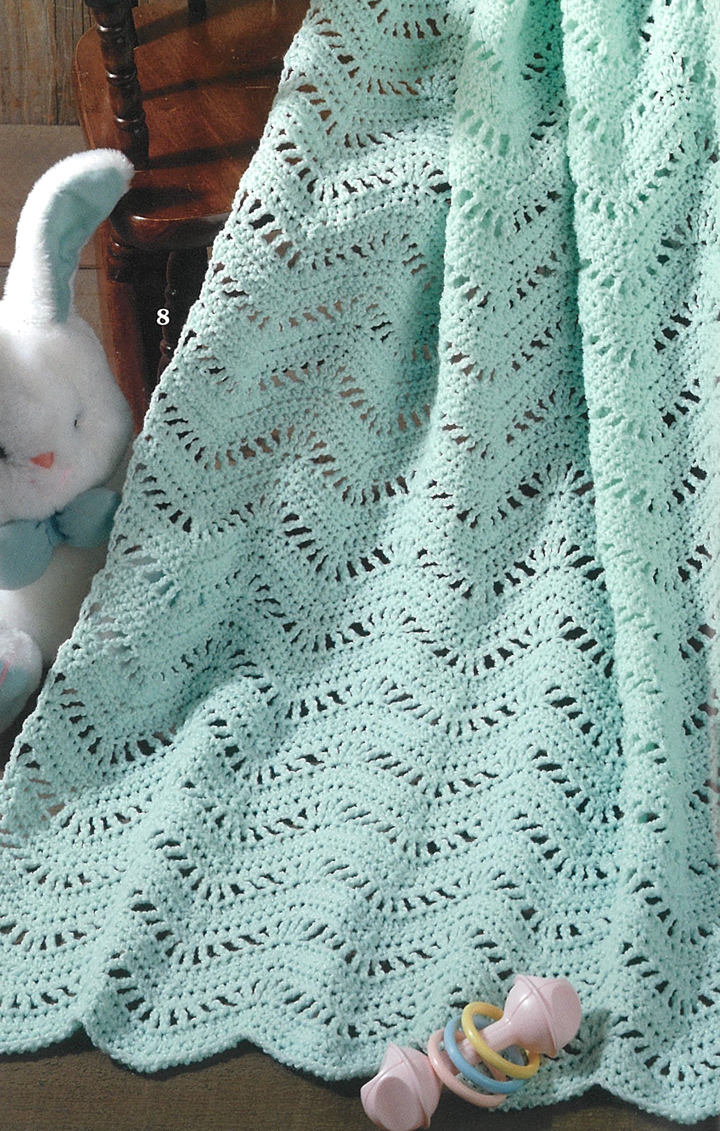 Leisure Arts' "Beginner's Guide Ripples For Baby" features a soft mint green chevron baby blanket—ideal for new crocheters—shown draped over a chair beside a white plush bunny and pink rattle on a brown surface.