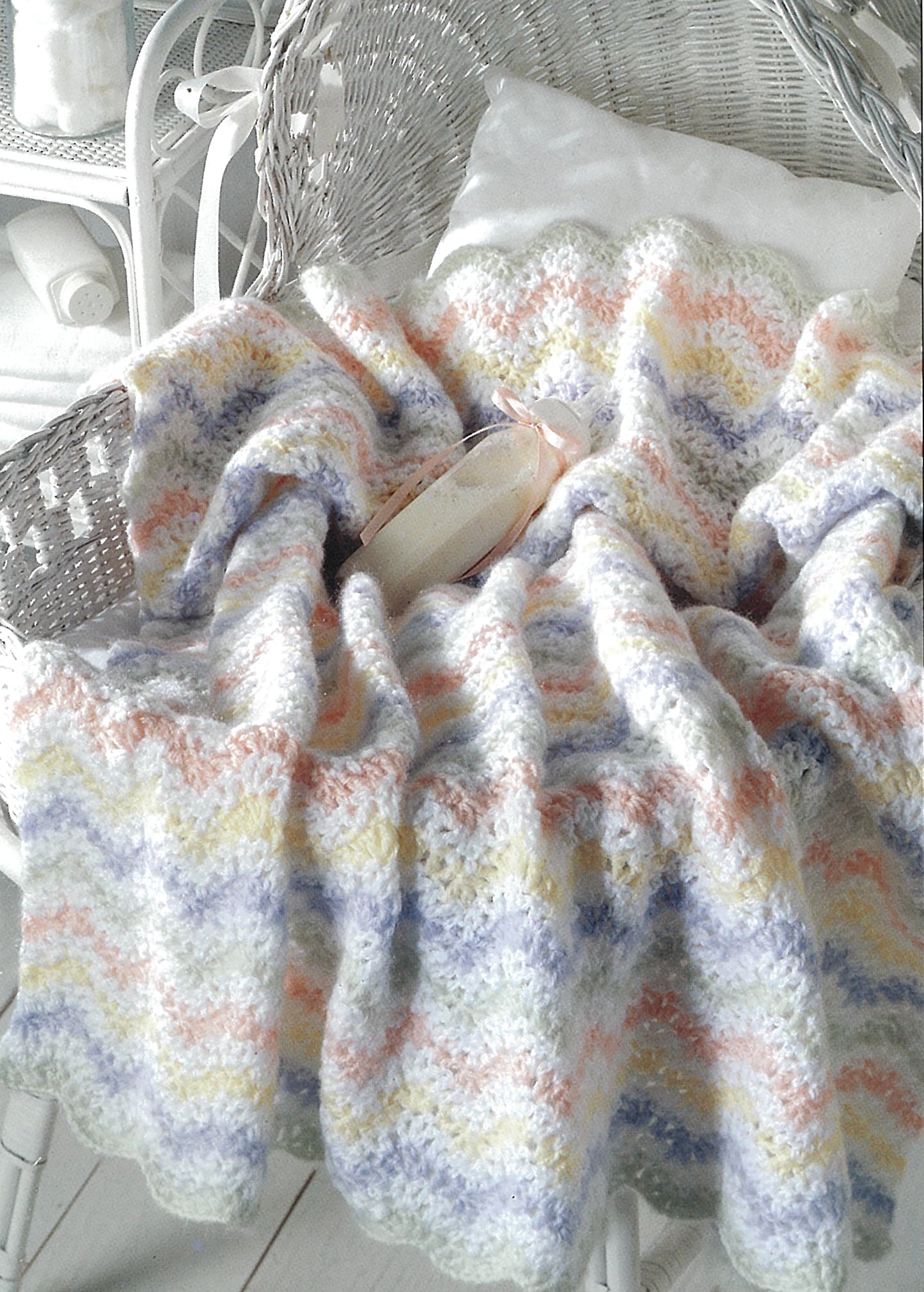 The Leisure Arts Beginner's Guide Ripples For Baby pastel, wavy-patterned crocheted blanket—great for beginners—is draped over a white wicker chair, with a white pillow and pink ballet slippers on top.