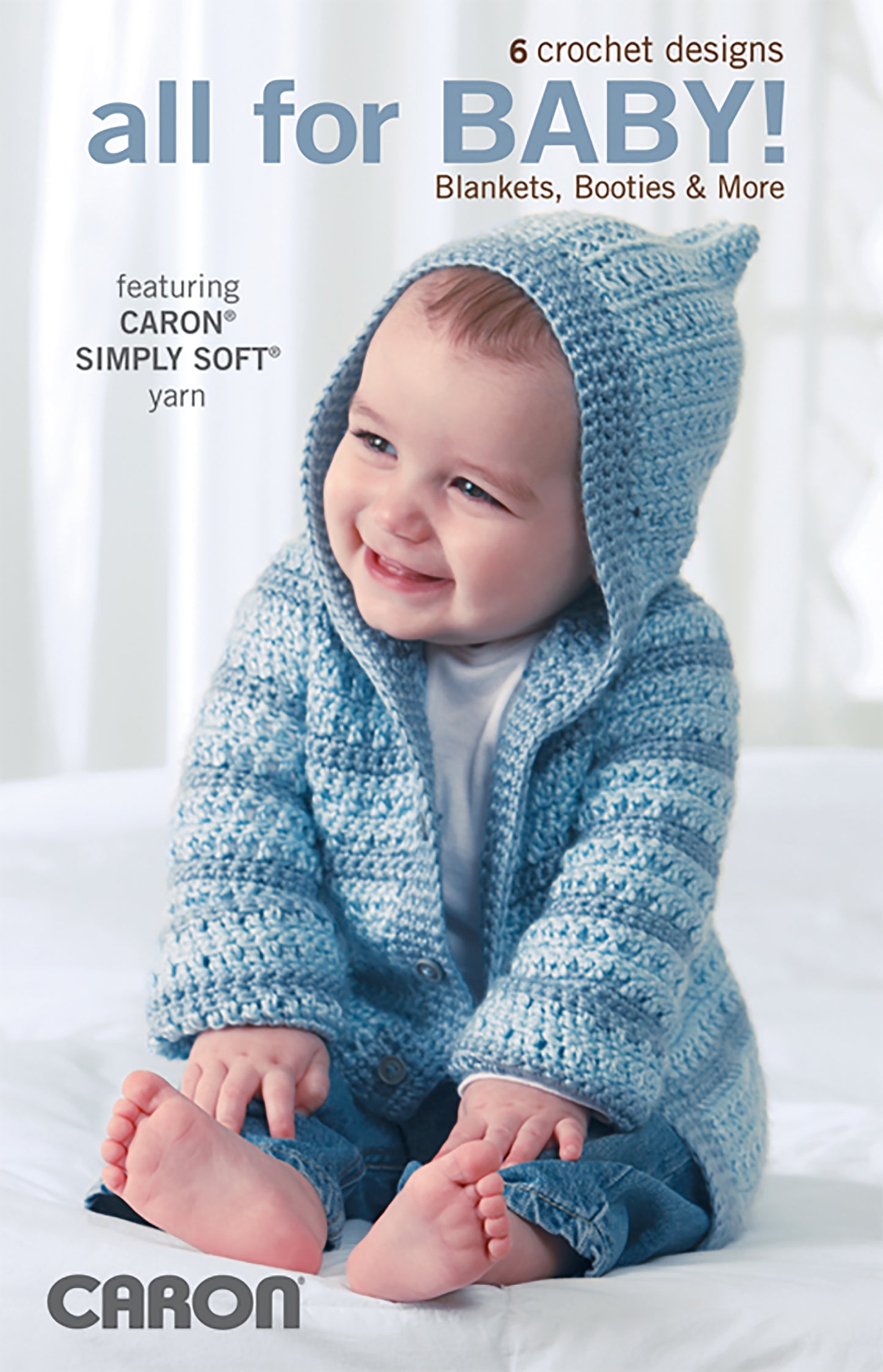 A smiling baby in a blue crocheted hoodie and booties, made with Caron Simply Soft yarn, sits on a white surface. The image promotes All For Baby!—6 Designs by Lori Markovic Digital Download from Leisure Arts.