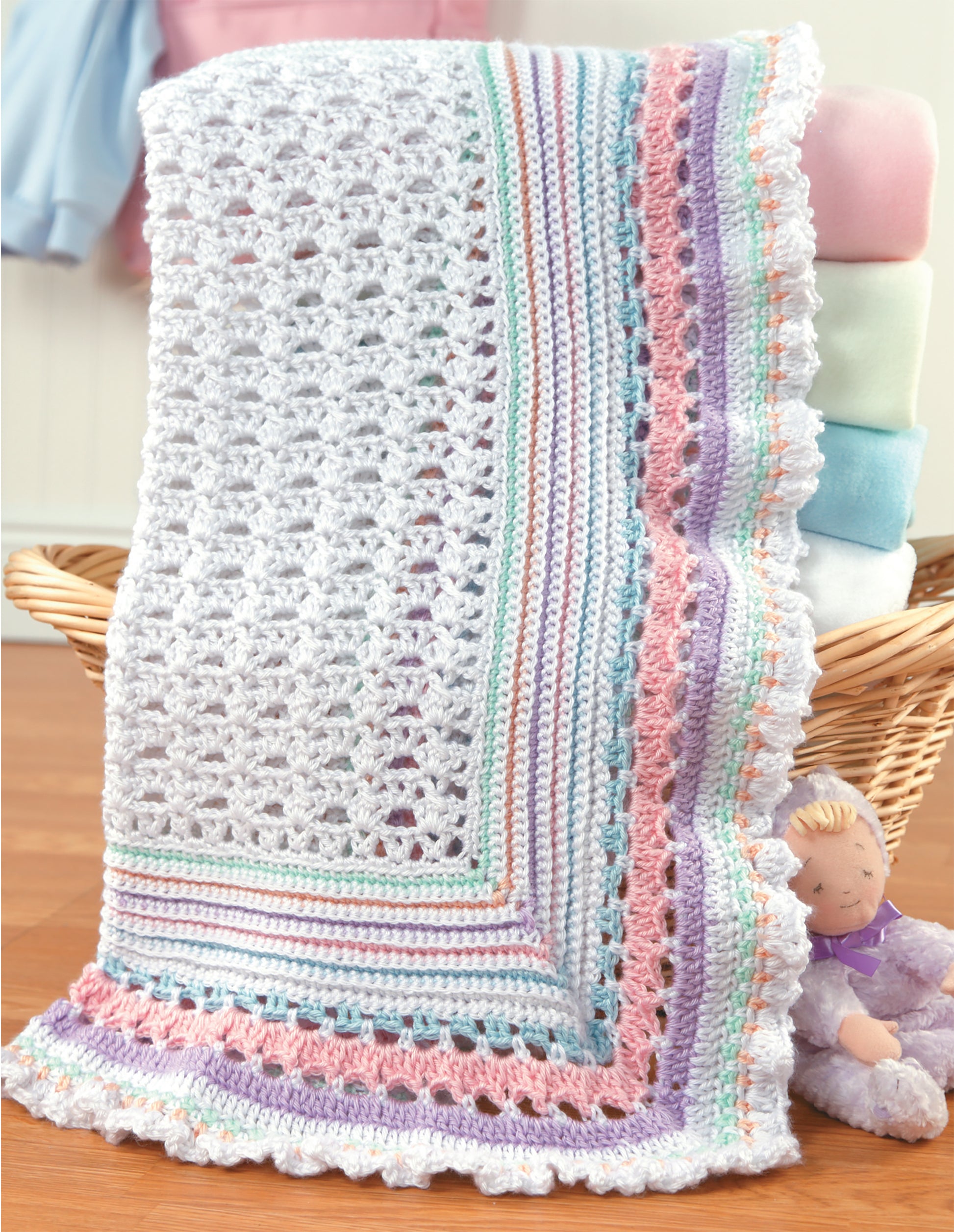A blanket made from the All For Baby!—6 Designs by Lori Markovic (Leisure Arts) digital pattern hangs over a basket of folded blankets, with a purple plush toy and baby booties beside it on a wooden floor.