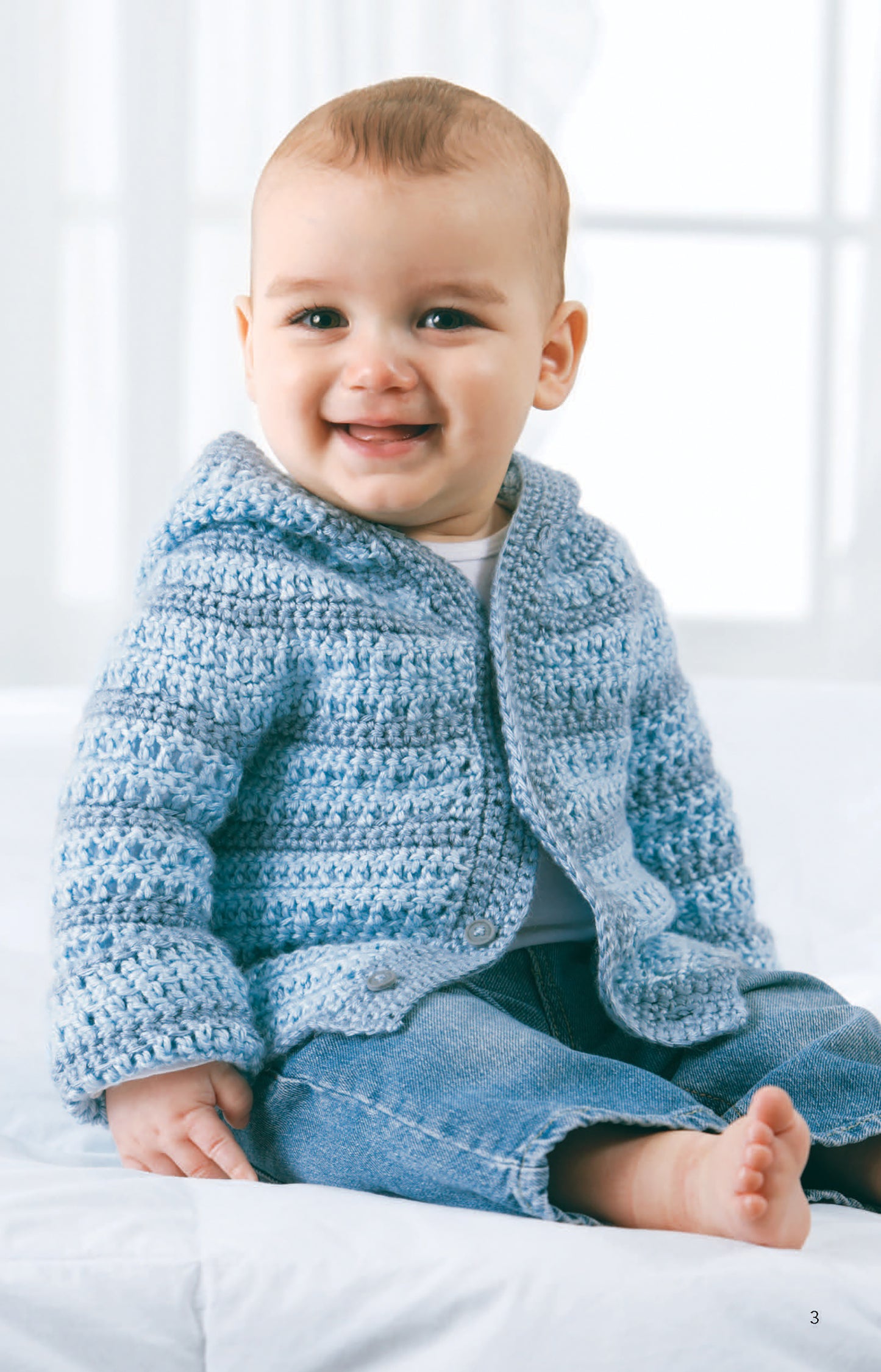 A smiling baby in a light blue Caron Simply Soft yarn hoodie and jeans sits barefoot on white, evoking the cozy charm of adorable crochet patterns from "All For Baby!—6 Designs by Lori Markovic" by Leisure Arts.