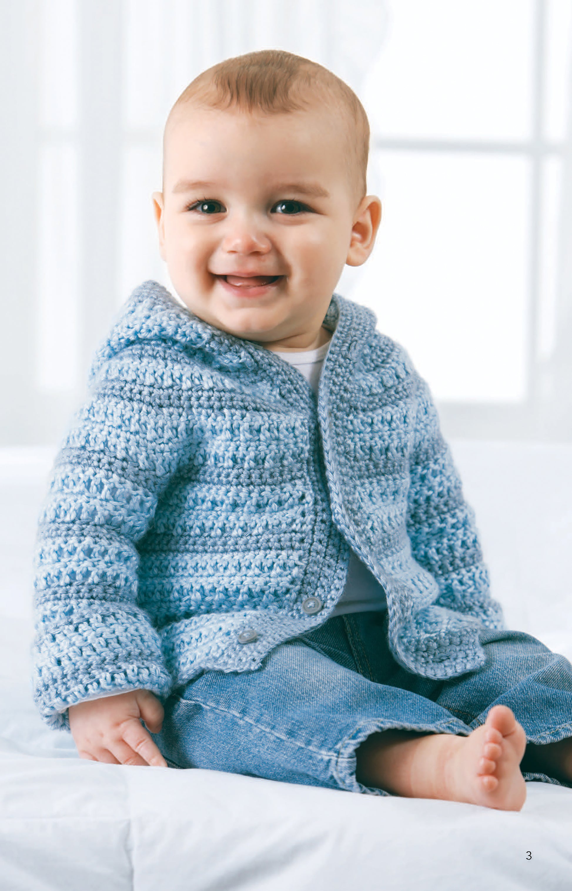 A smiling baby in a light blue Caron Simply Soft yarn hoodie and jeans sits barefoot on white, evoking the cozy charm of adorable crochet patterns from "All For Baby!—6 Designs by Lori Markovic" by Leisure Arts.