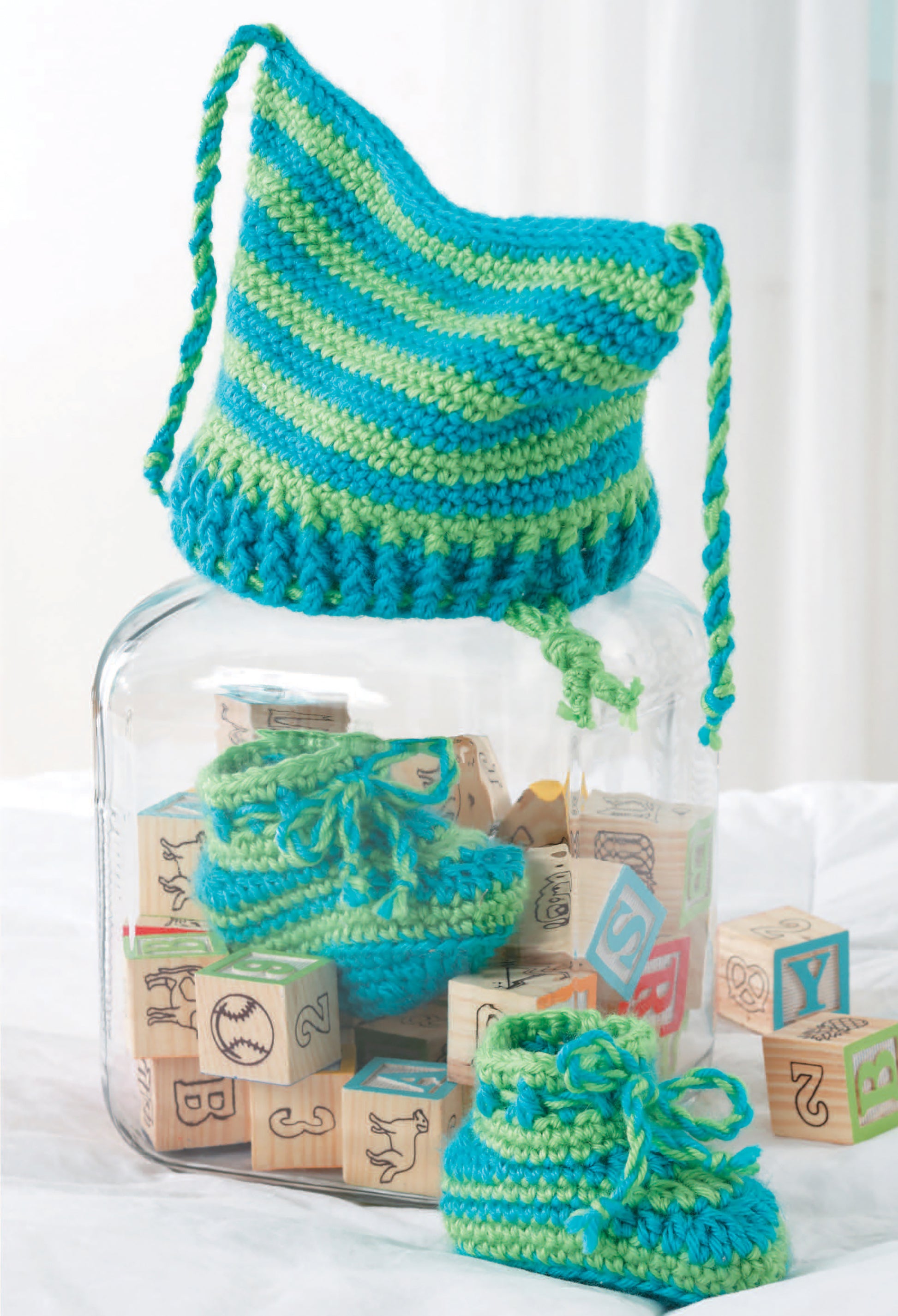 A blue and green striped hat with matching booties, made using Caron Simply Soft yarn, is displayed with a glass jar of wooden blocks. Featured in "All For Baby!—6 Designs by Lori Markovic" (Leisure Arts, digital download). White blurred background.