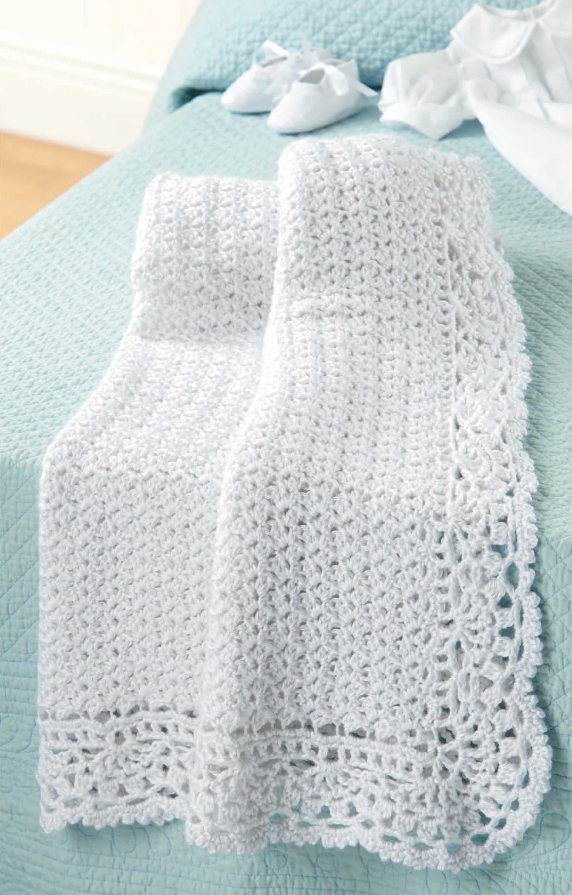 A white crocheted baby blanket made with Caron Simply Soft yarn, shown on a blue quilted bed with baby booties nearby, features in "All For Baby!—6 Designs by Lori Markovic," available as a Leisure Arts digital download.