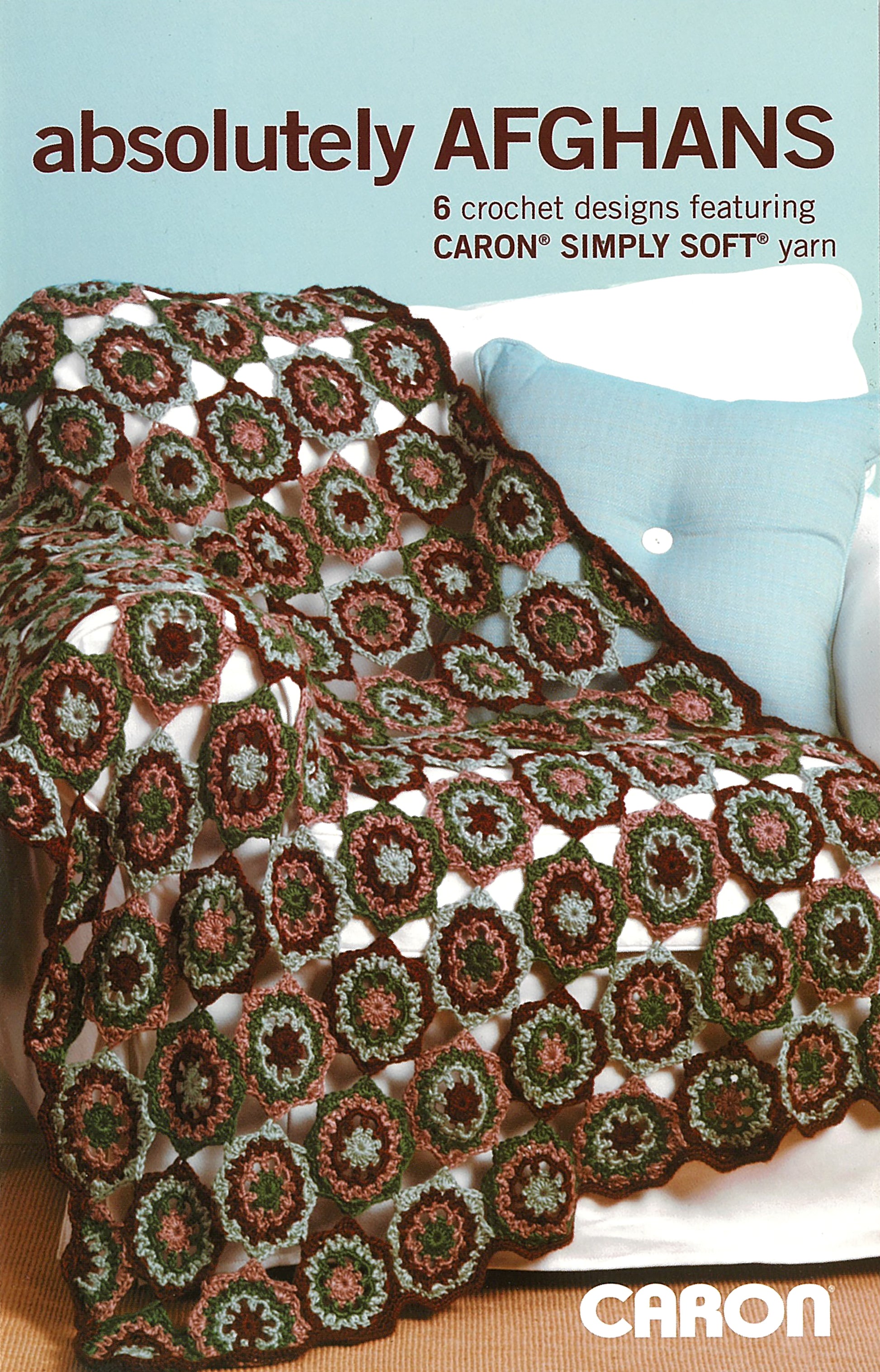A cozy afghan with circular and granny square motifs in earth tones decorates a white chair with a blue pillow. Text: “Absolutely Afghans Crochet by Leisure Arts—6 crochet designs using Caron Simply Soft yarn.”.