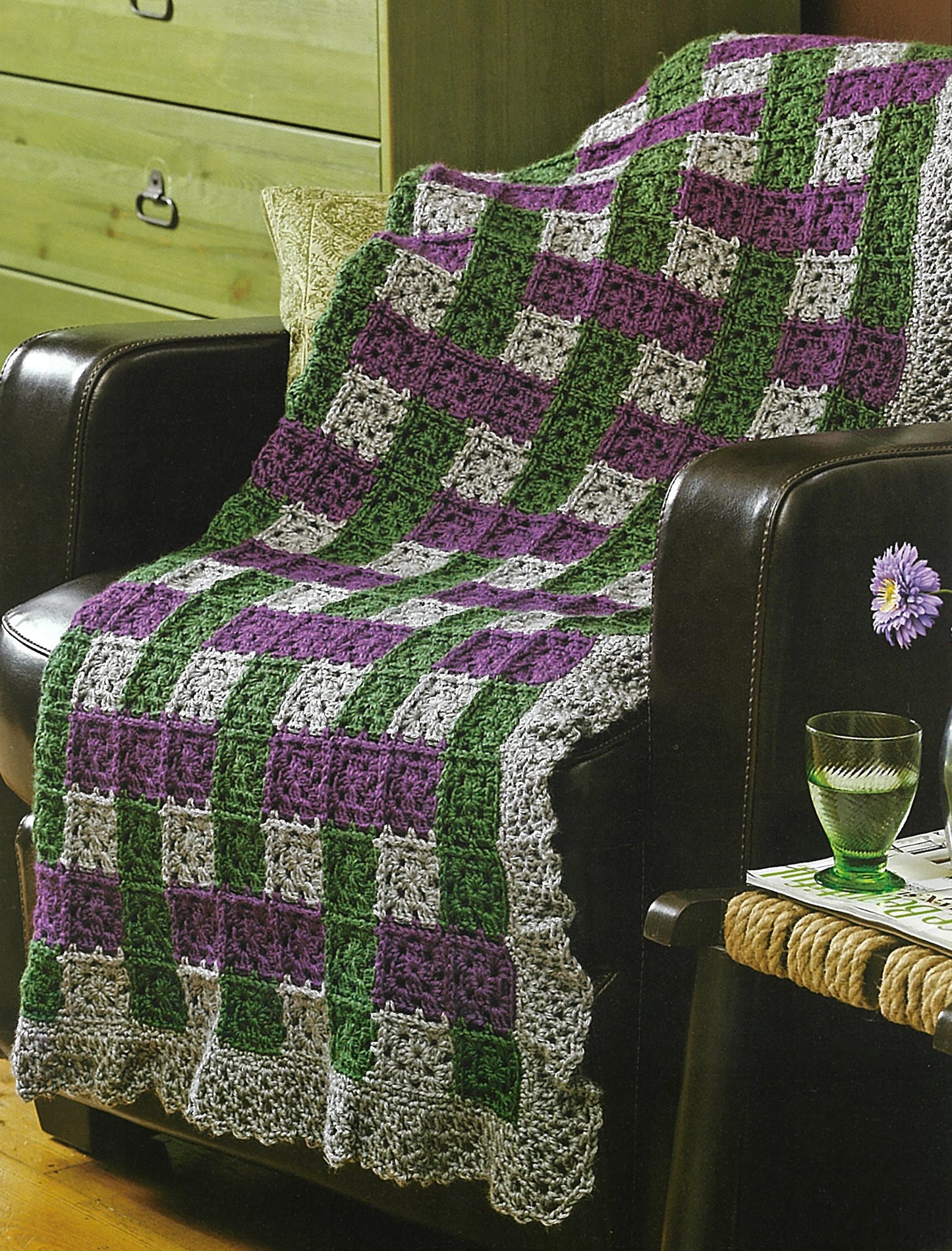 An Absolutely Afghans Crochet blanket by Leisure Arts, featuring a Caron Simply Soft yarn checkerboard in purple, green, and gray, adorns a dark armchair next to a wooden dresser and a table with a flower, glass, and book.