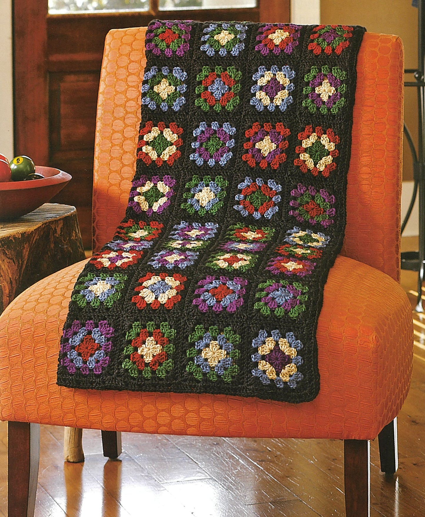 A vibrant granny square blanket from Absolutely Afghans Crochet by Leisure Arts is draped over an orange textured chair in a cozy room with a wooden floor and a bowl of fruit on the nearby table.