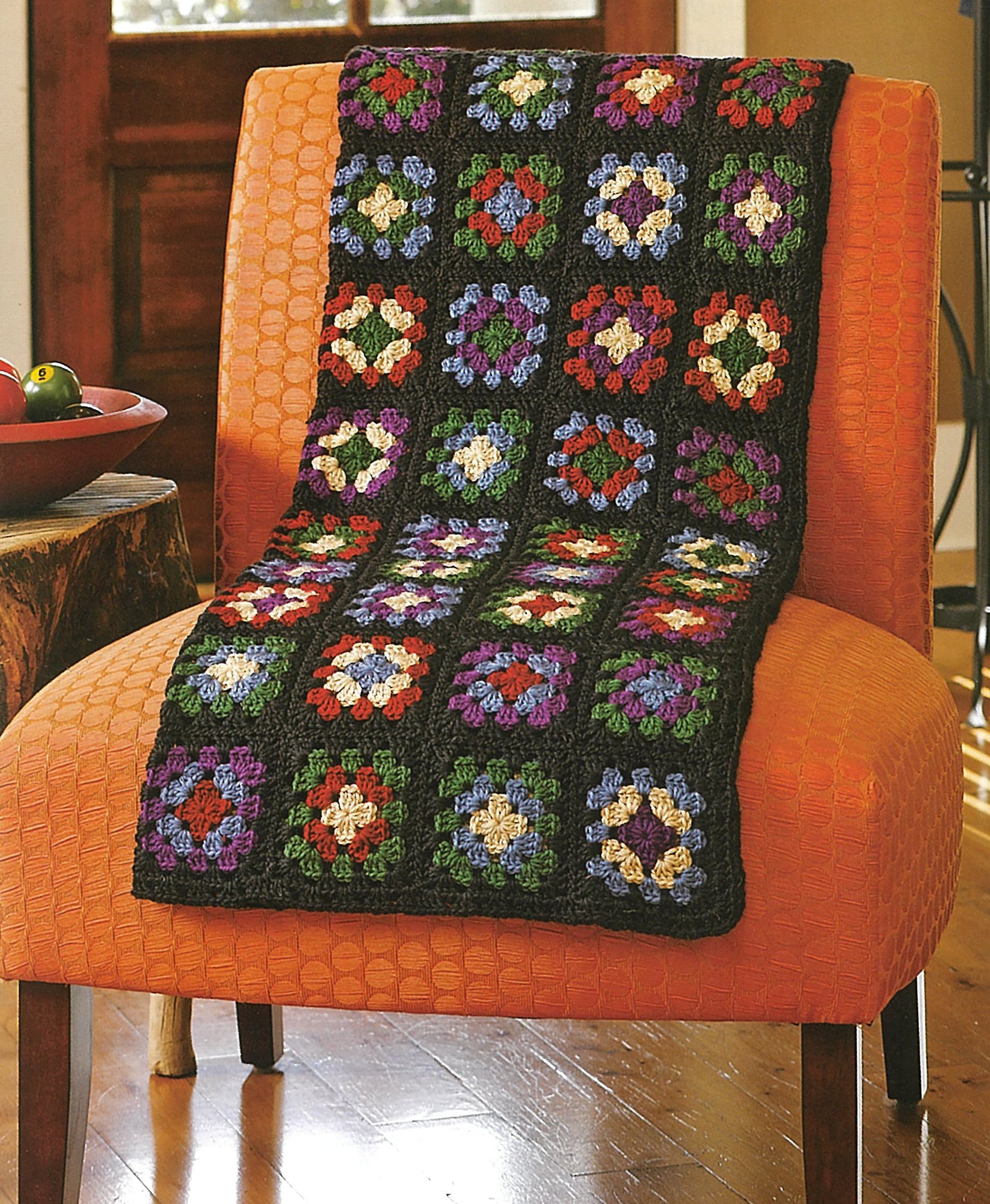 A vibrant granny square blanket from Absolutely Afghans Crochet by Leisure Arts is draped over an orange textured chair in a cozy room with a wooden floor and a bowl of fruit on the nearby table.