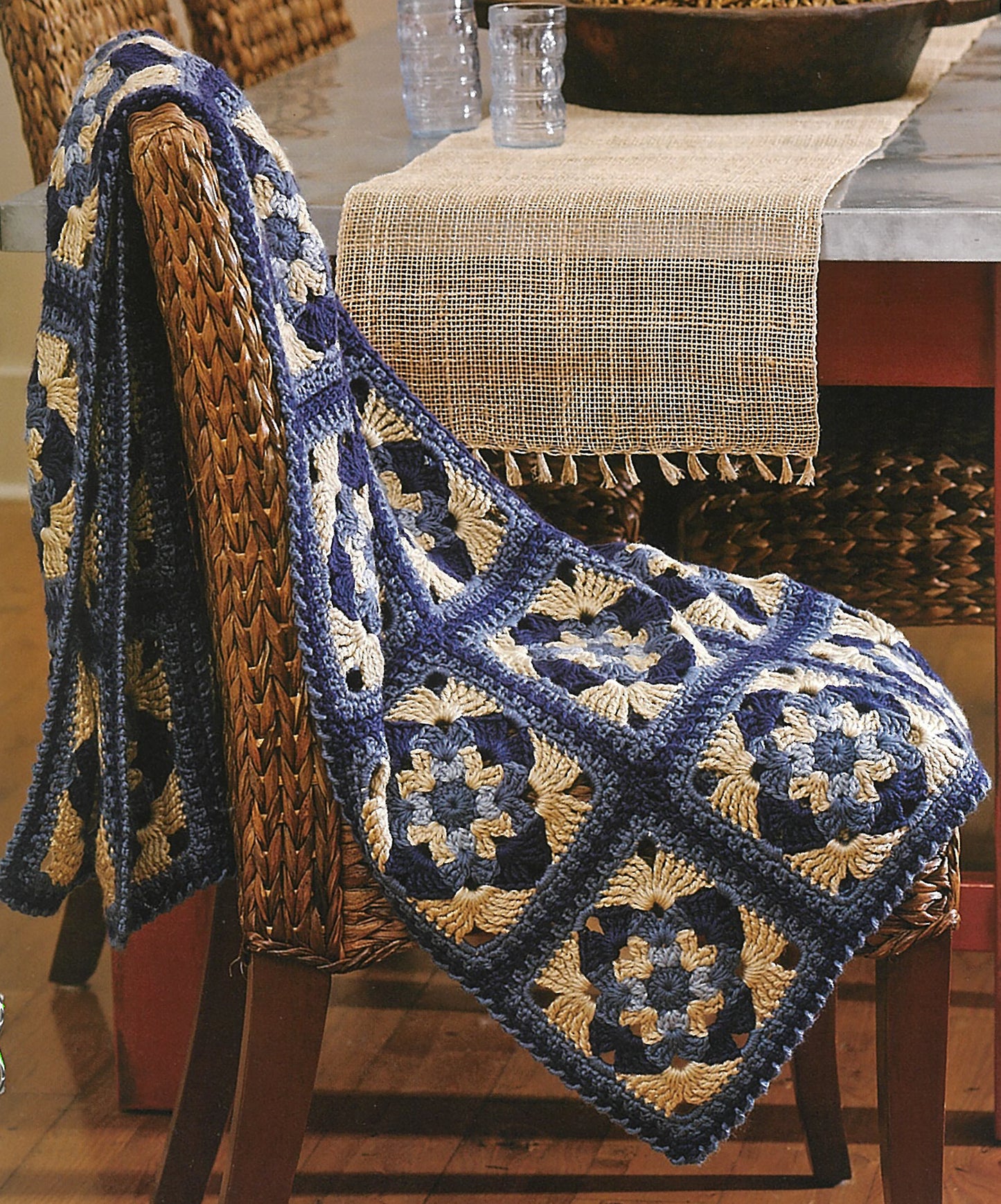 The "Absolutely Afghans Crochet" blanket by Leisure Arts, featuring a blue, cream, and beige granny square pattern, is draped over a woven chair in a cozy dining room.
