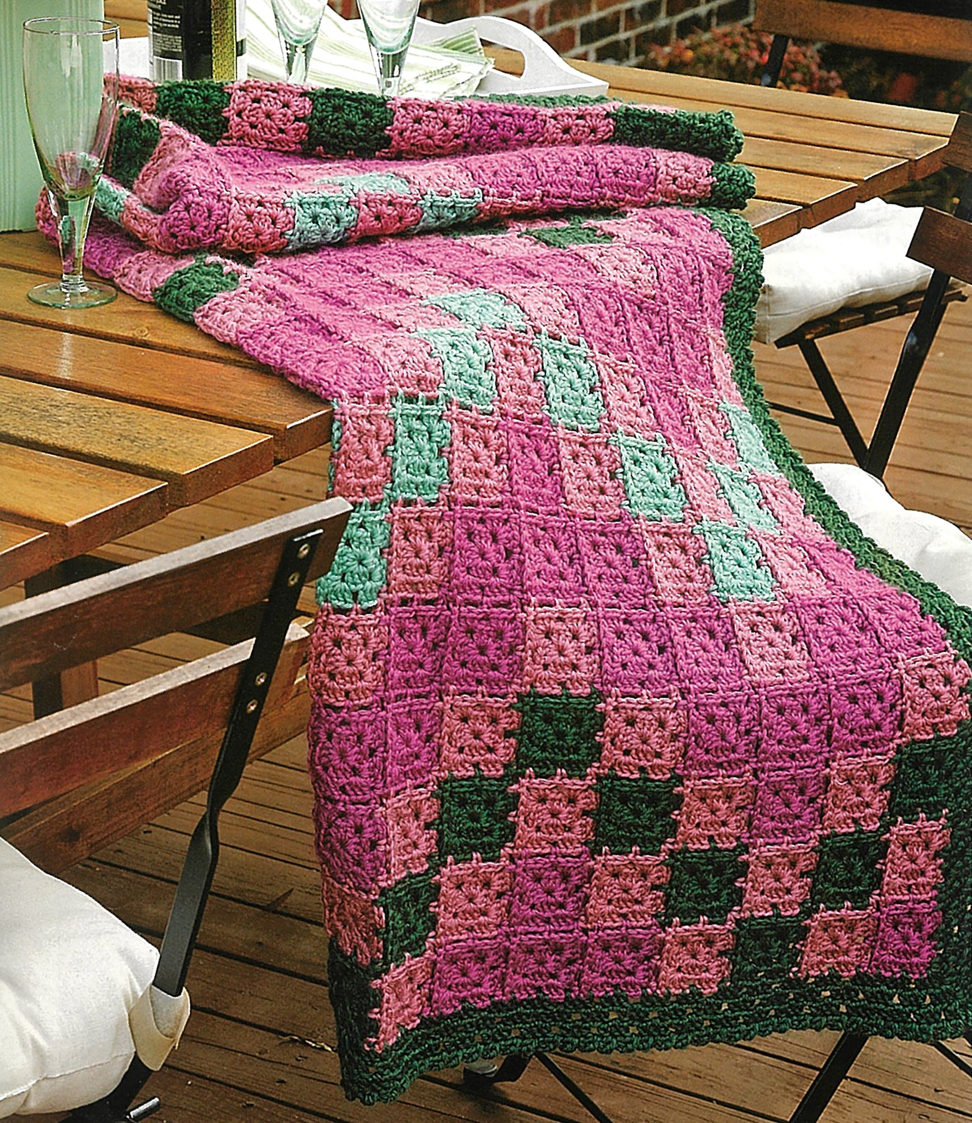 A vibrant crocheted blanket from Leisure Arts' Absolutely Afghans Crochet drapes over a wooden outdoor table, showcasing pink, green, and purple granny squares beside chairs and glassware.