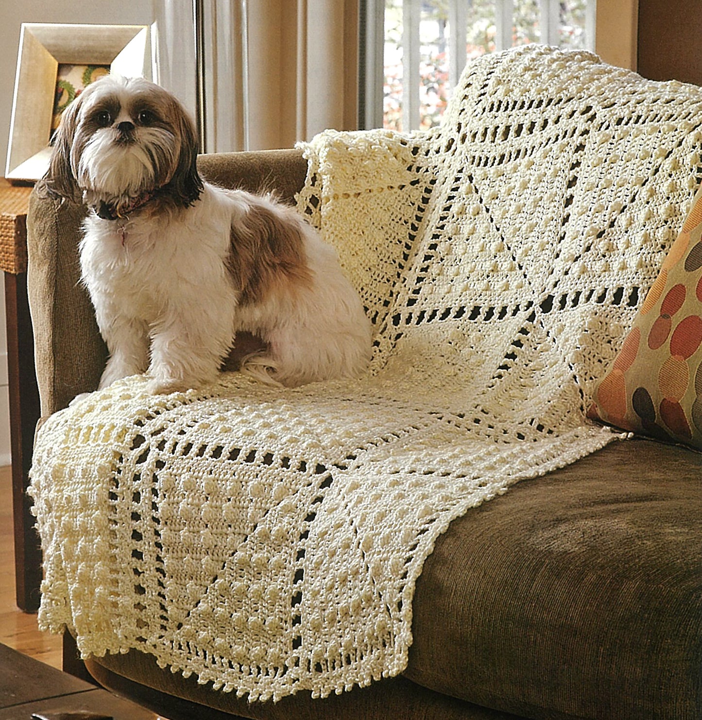 A small, fluffy dog with white and brown fur sits on a brown couch draped in a cream granny square blanket made using the Absolutely Afghans Crochet book by Leisure Arts. A patterned pillow and a sunlit window are in the background.