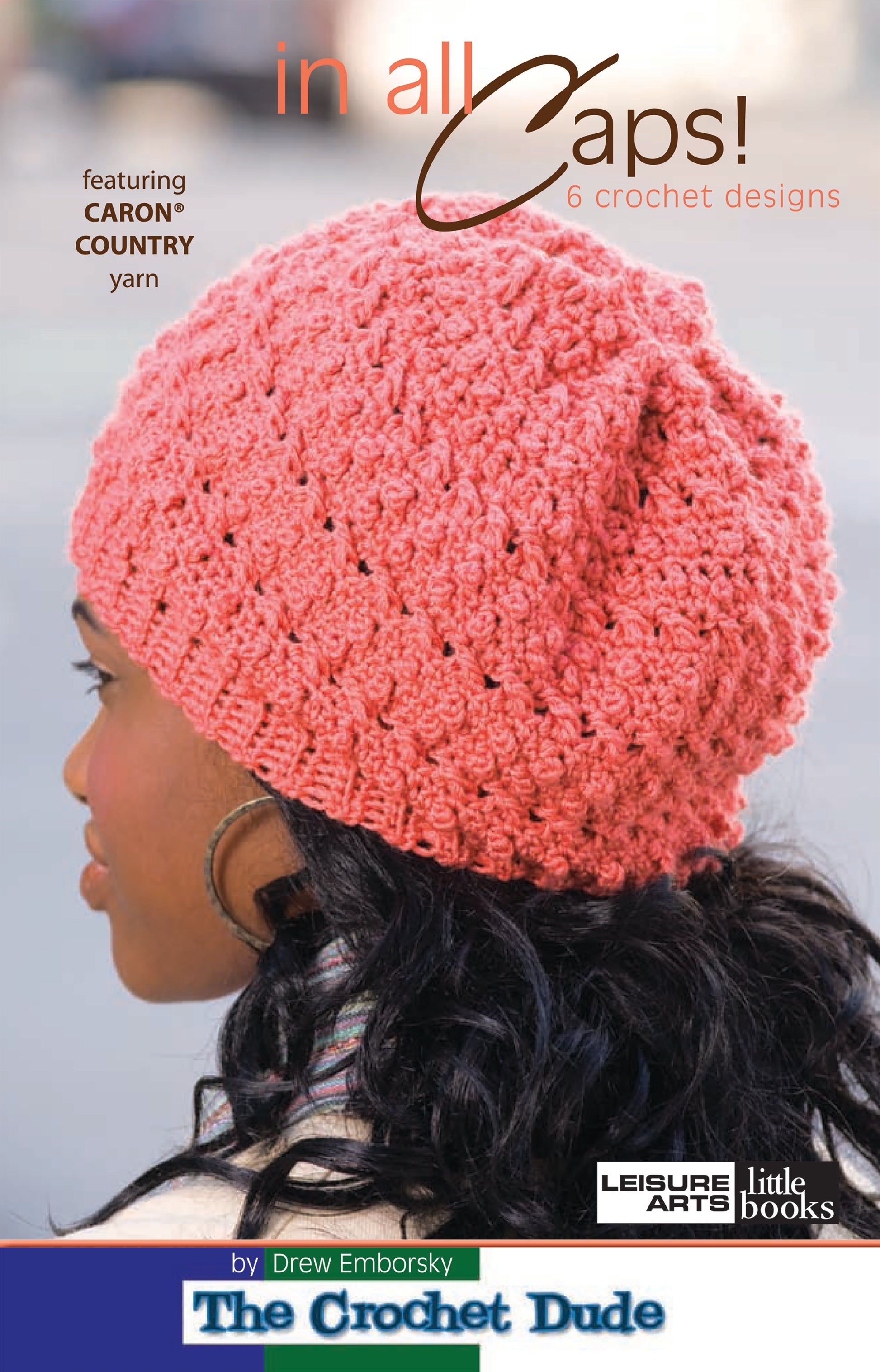 A woman with long dark hair wears a textured pink crochet hat. The cover text reads: In All Caps! 6 crochet designs featuring medium weight Caron Country Yarn, by The Crochet Dude Drew Emborsky. Brand: Leisure Arts.