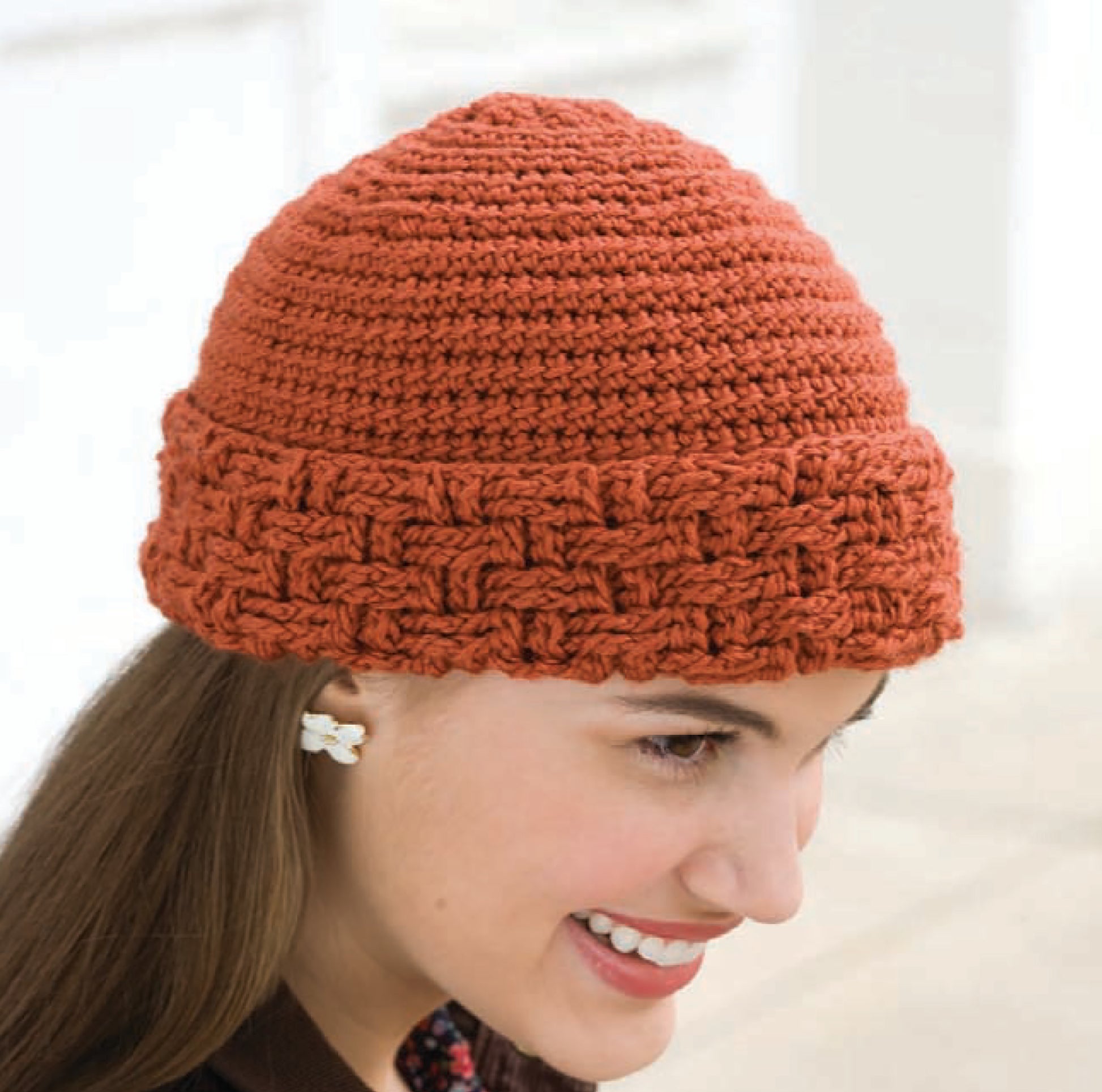 A smiling person wears the "In All Caps!" beanie by Leisure Arts—a rust-colored, textured hat made from medium-weight yarn with a thick patterned brim—paired with a small white flower-shaped earring.