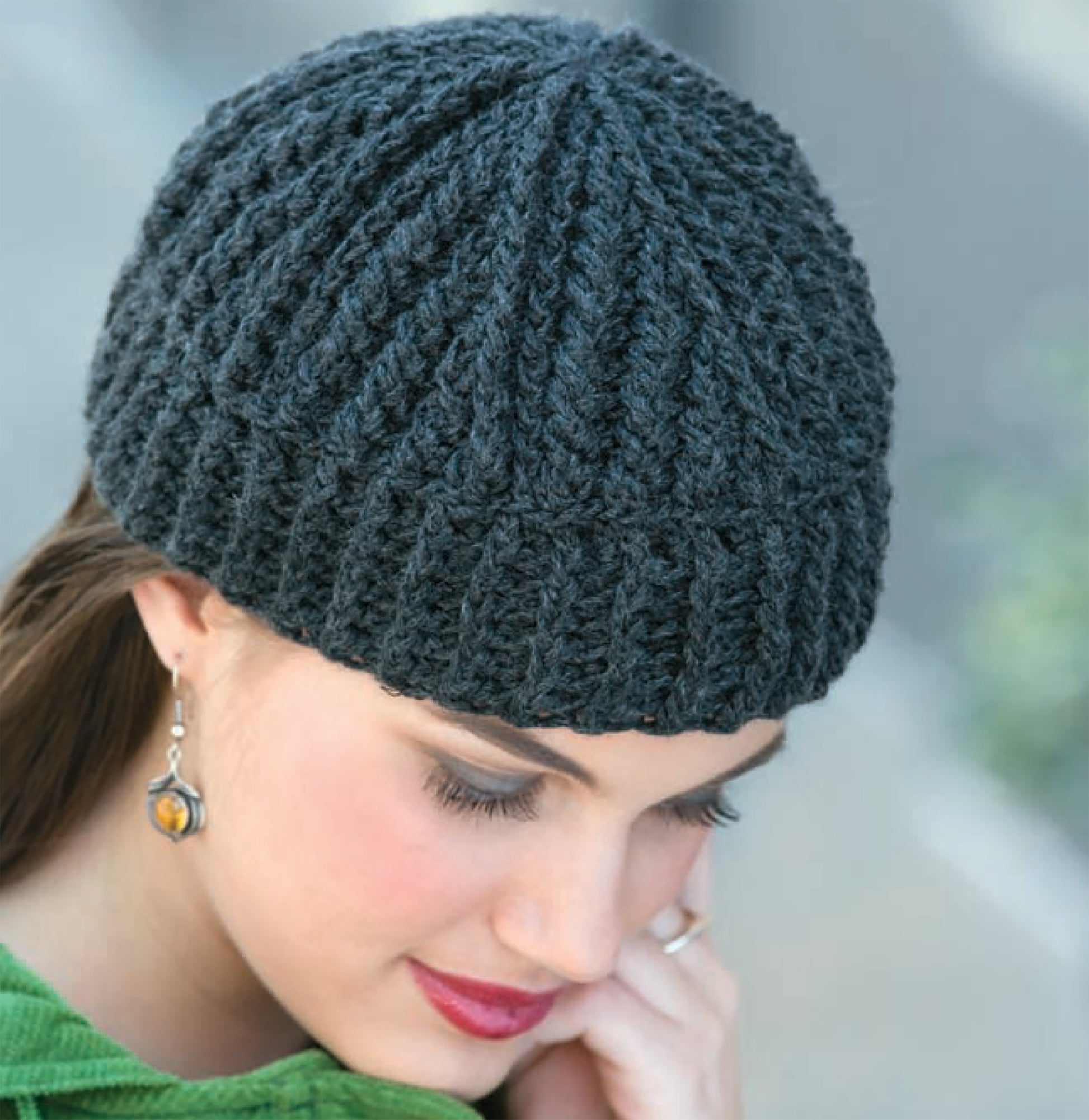 A woman wearing the "In All Caps!" dark gray knitted beanie by Leisure Arts looks downward. She has long brown hair, an amber stone earring, and a green top.