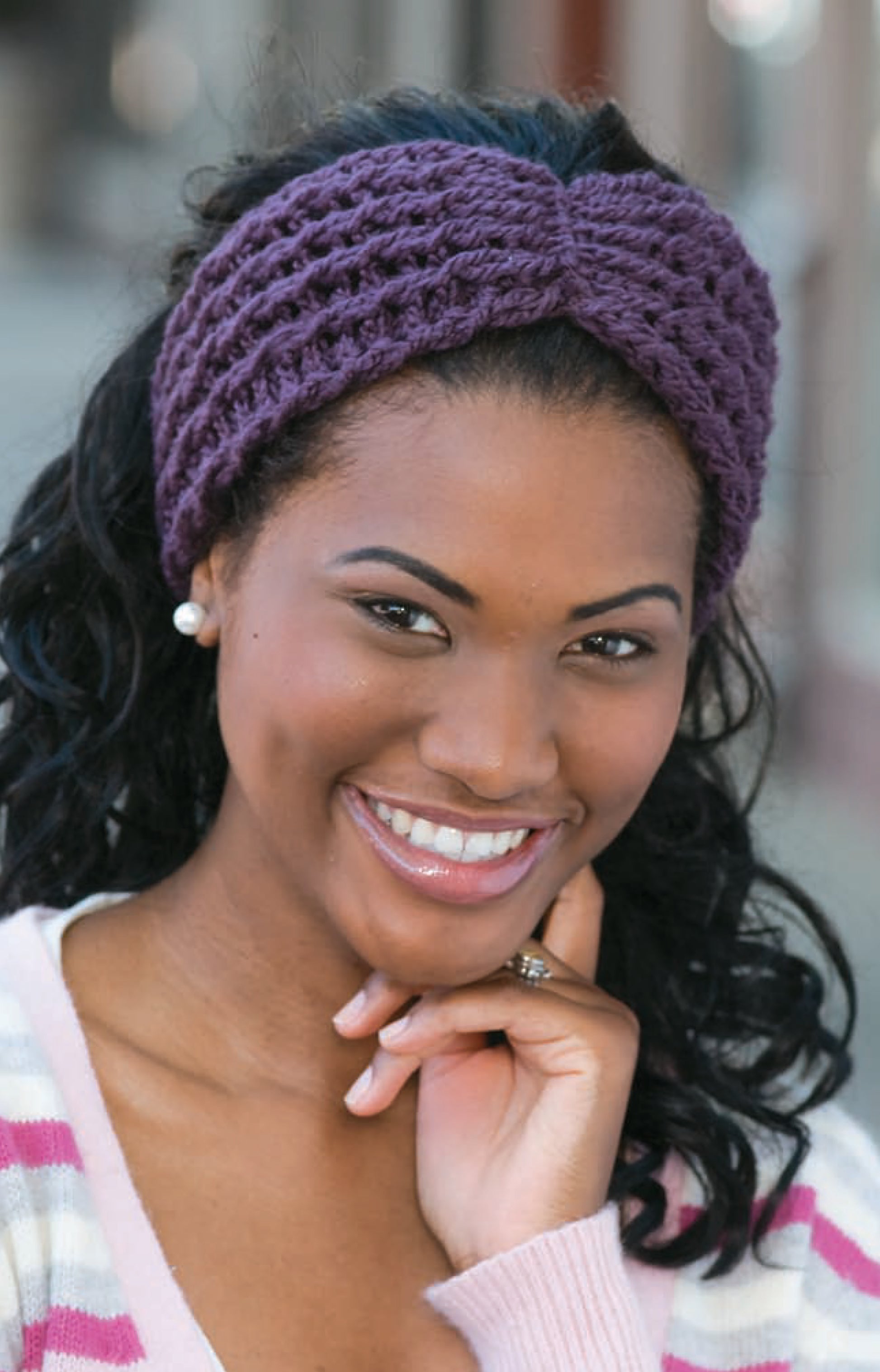 A woman with long dark hair wears the "In All Caps!" headband by Leisure Arts, made from medium weight yarn. She smiles, resting her chin on her hand outdoors, styled with pearl earrings and a striped sweater.