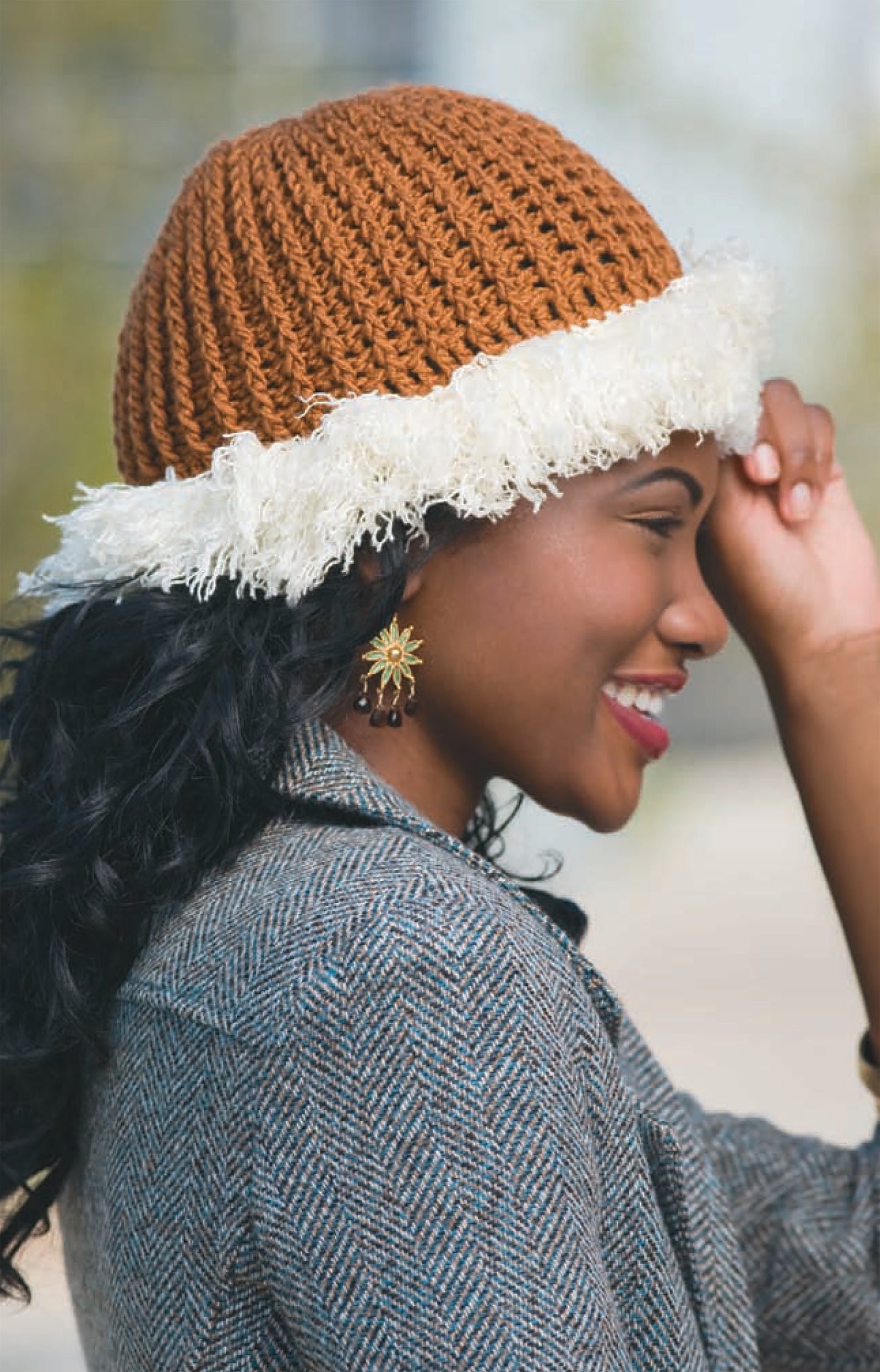 A woman smiles outdoors in a gray textured coat and sunburst earrings, holding the brim of her brown "In All Caps!" hat by Leisure Arts—crafted with medium-weight yarn and fluffy white trim.