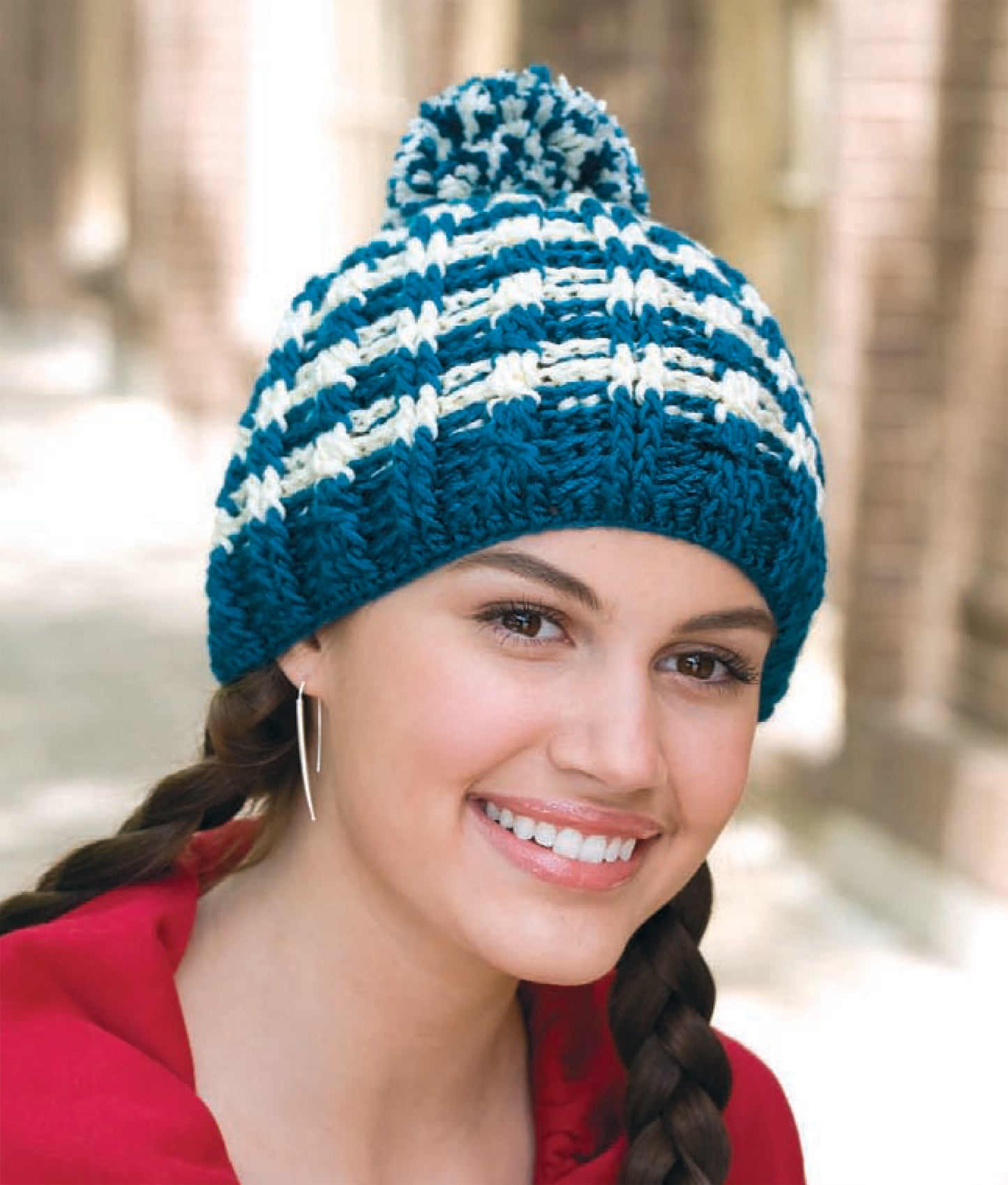 A smiling person with braided hair wears the Leisure Arts "In All Caps!" blue and white striped knit hat with a pom-pom, paired with a red hoodie, standing outdoors by a brick building.