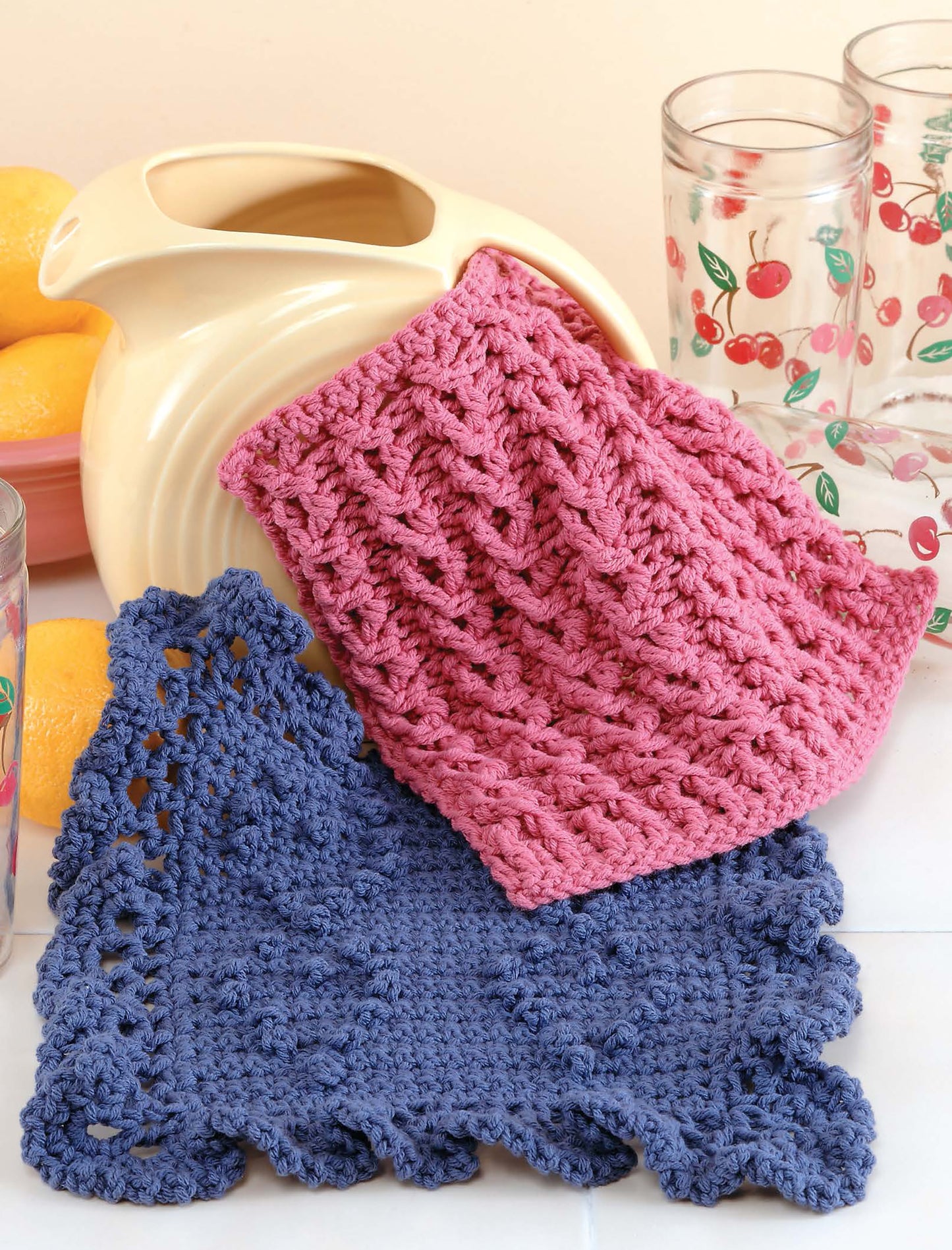 Displayed on a white surface are two crochet dishcloths—one pink, one blue—beside a yellow pitcher, cherry glasses, and lemons. Get inspired to create your own with "All Washed Up—9 Crochet Dishcloths" by Leisure Arts.