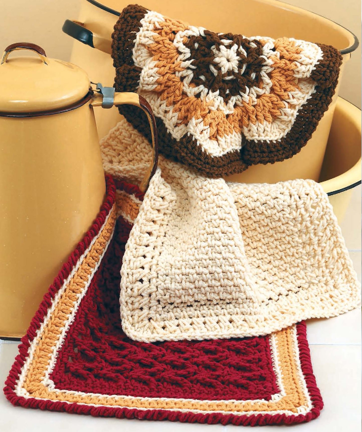 Leisure Arts' All Washed Up—9 Crochet Dishcloths are shown in cream, brown, gold, and red hues draped over a yellow enamel pot. These textured DIY dishcloths feature contrasting borders for a handcrafted appeal.