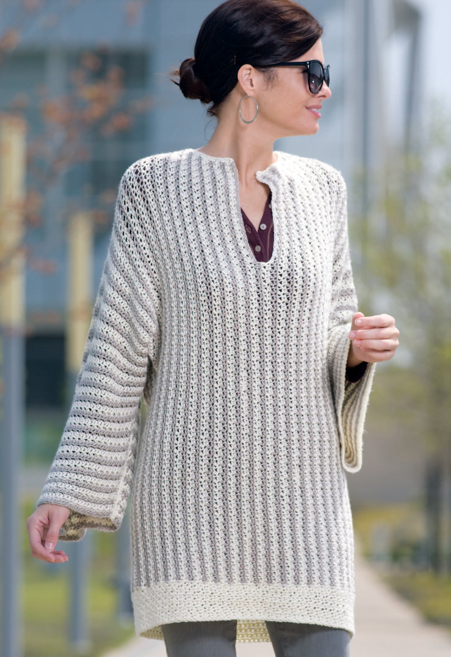 A woman outdoors in sunglasses and hoop earrings wears a cream and gray striped knit sweater inspired by Relaxed Fashion—4 Crochet Designs Featuring Caron Spa Yarn from Leisure Arts, walking on a sunny day.